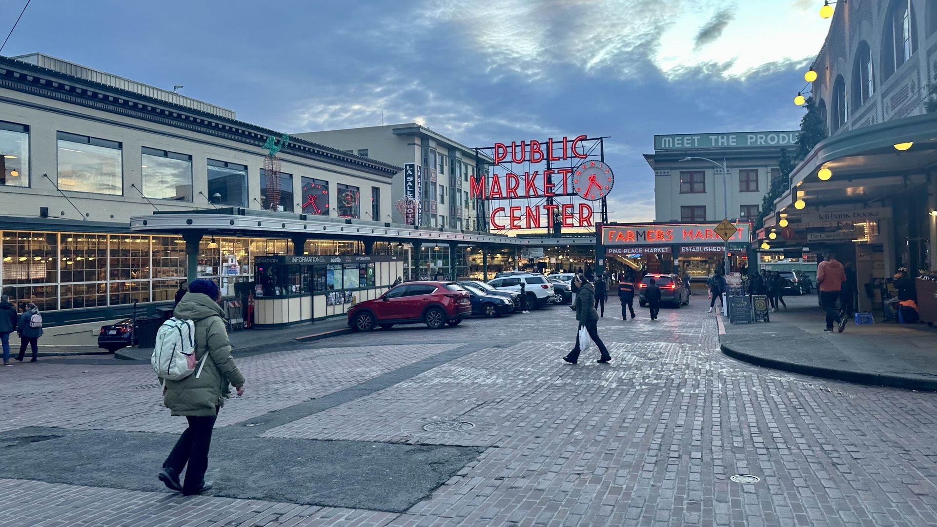 People walk in front of Pike Place Market and its neon sign that says "Public Market Center" at twilight.