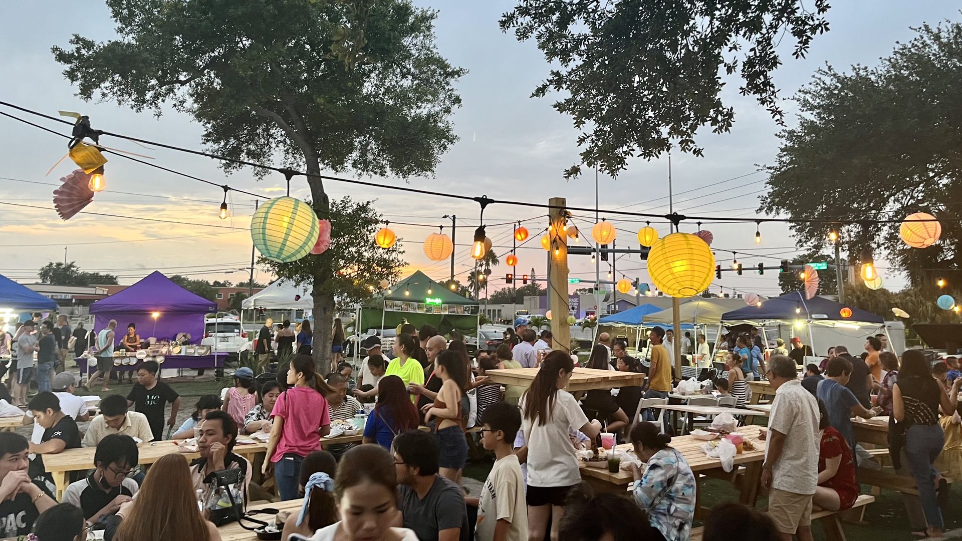 Paper lanterns hang over children and families eating at picnic tables.