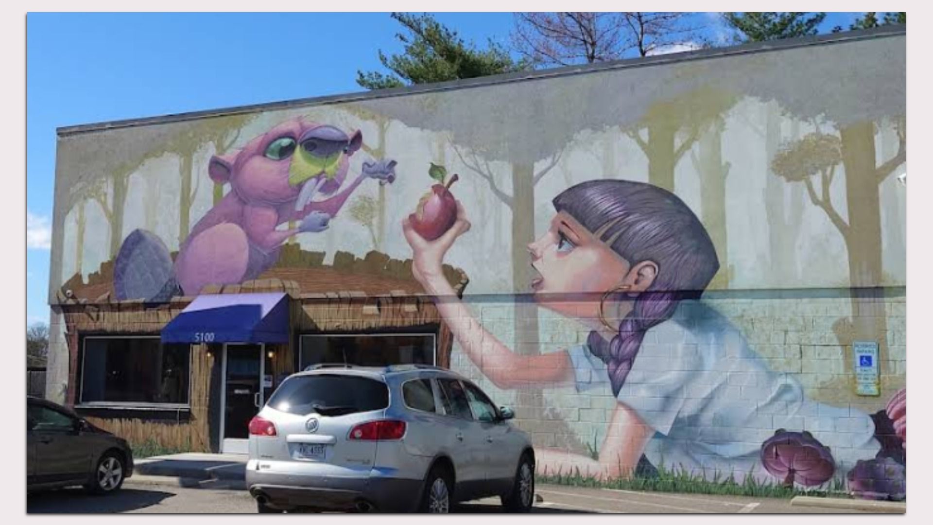 Mural on a building shows a girl with purple hair reaching toward a red apple offered by a pink creature in a forest scene; cars parked in front and a storefront with a blue awning.
