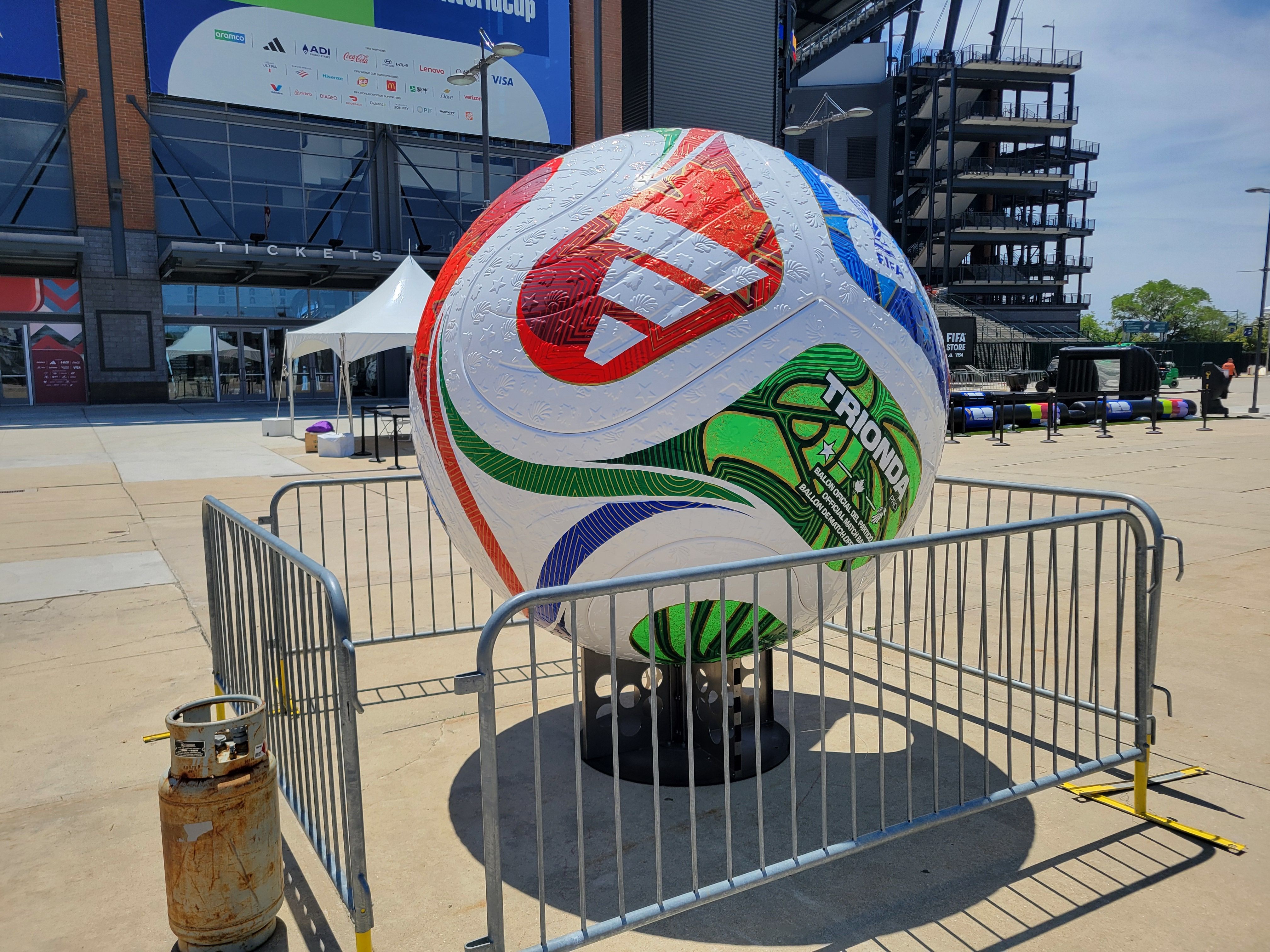 Colorful soccer ball sculpture with red, green, and blue panels sits behind a metal barrier outside a stadium; a TICKETS sign and scaffolding building are visible in the background.