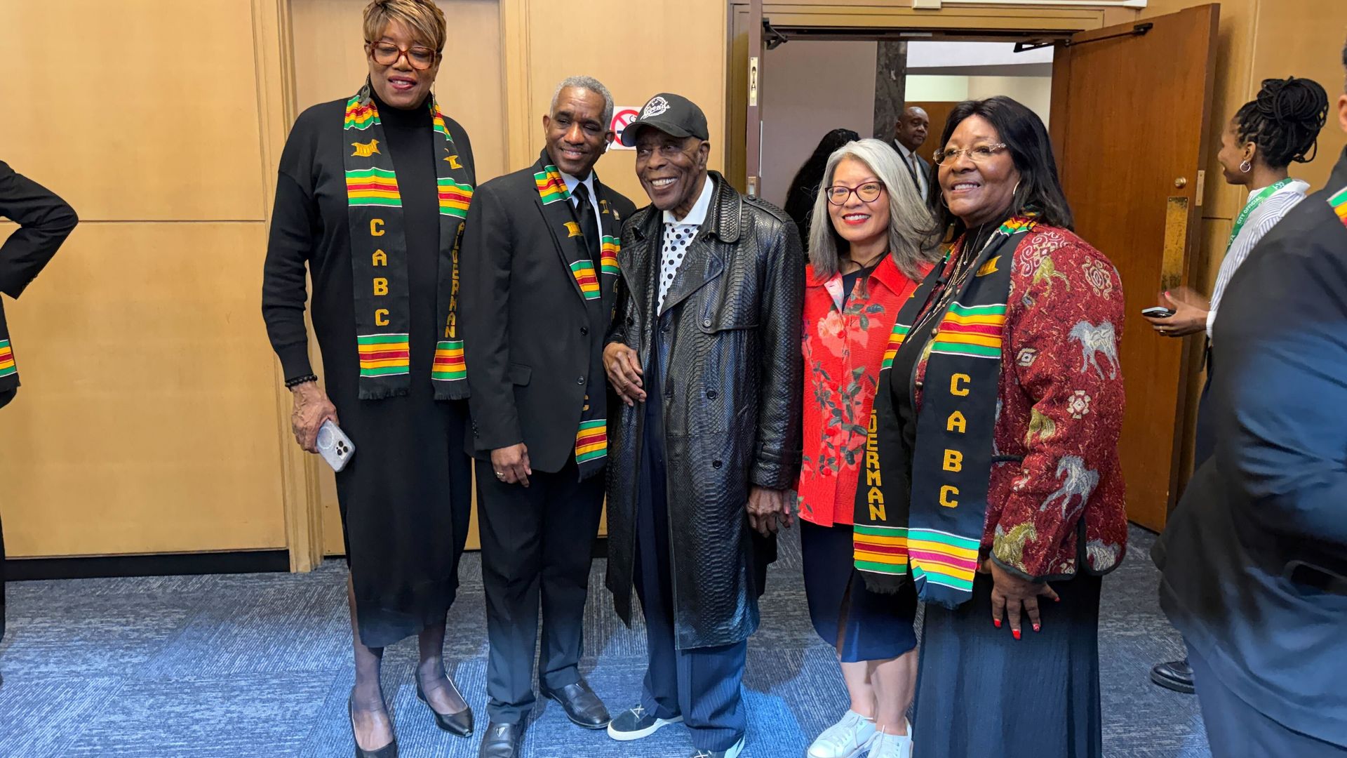 Group of five people standing indoors, four wearing colorful CABC kente stoles, smiling for a photo. One man in the center wears a black leather coat and cap; warm, friendly atmosphere.