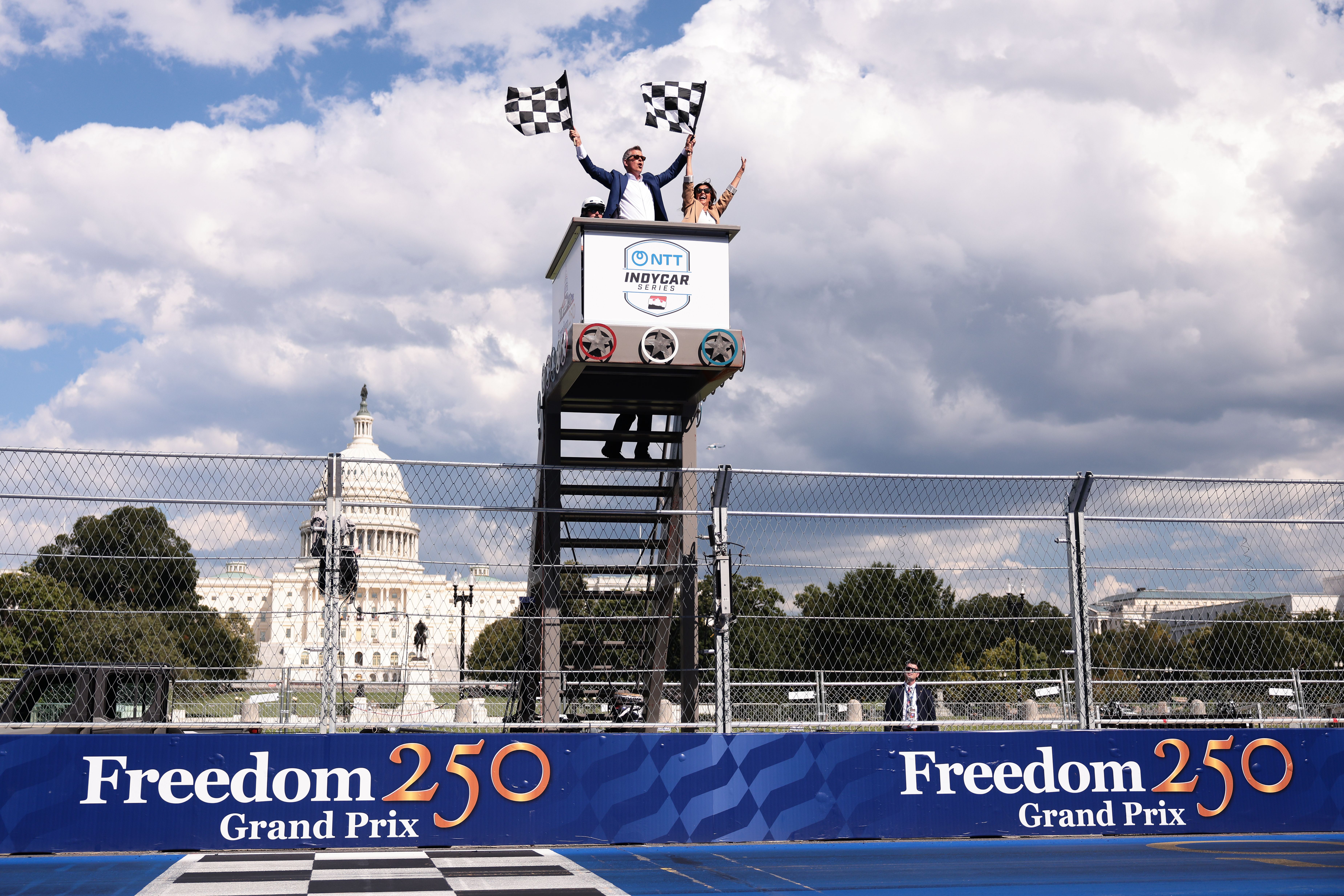 Transportation Secretary Sean Duffy waves the checkered flag on a podium with his wife Rachel Campos-Duffy in front of the Capitol.