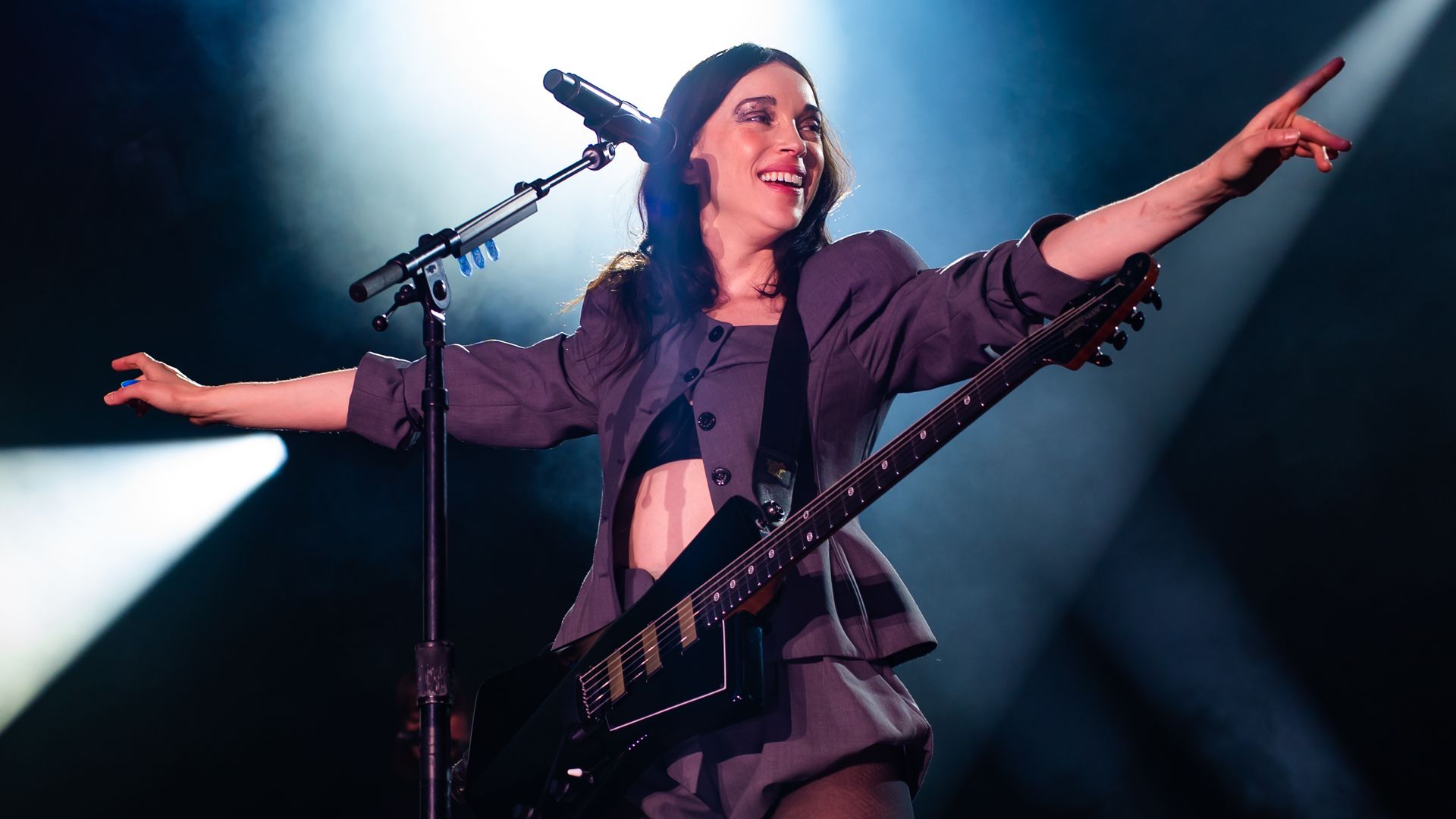 Smiling female musician in grey outfit with black electric guitar performs on stage under bright white spotlights, arms outstretched, standing behind a microphone.