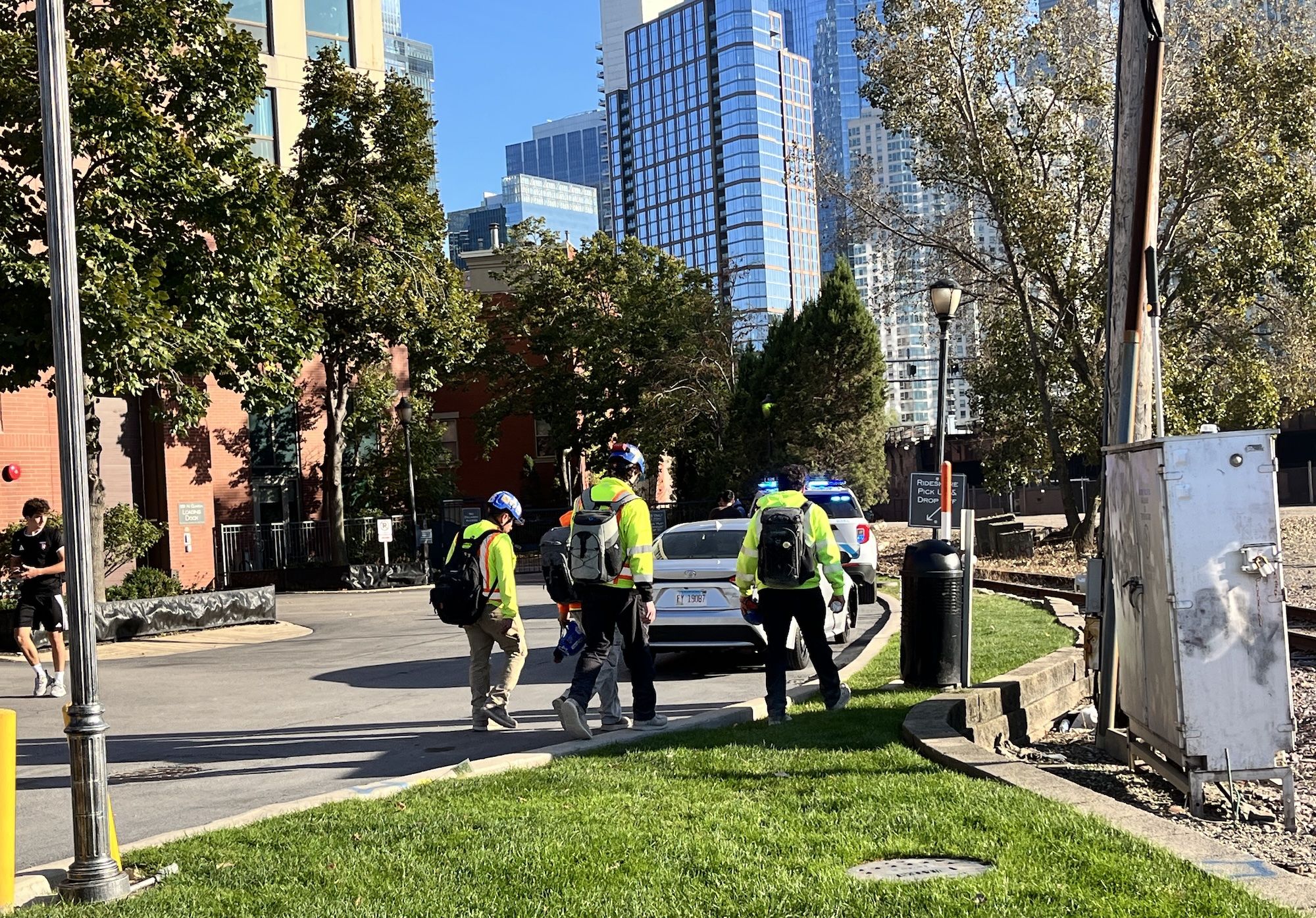 City street scene with tall glass buildings, leafy trees, and a bright blue sky. In the foreground, several people in neon safety vests and helmets stand near a police car with flashing lights.