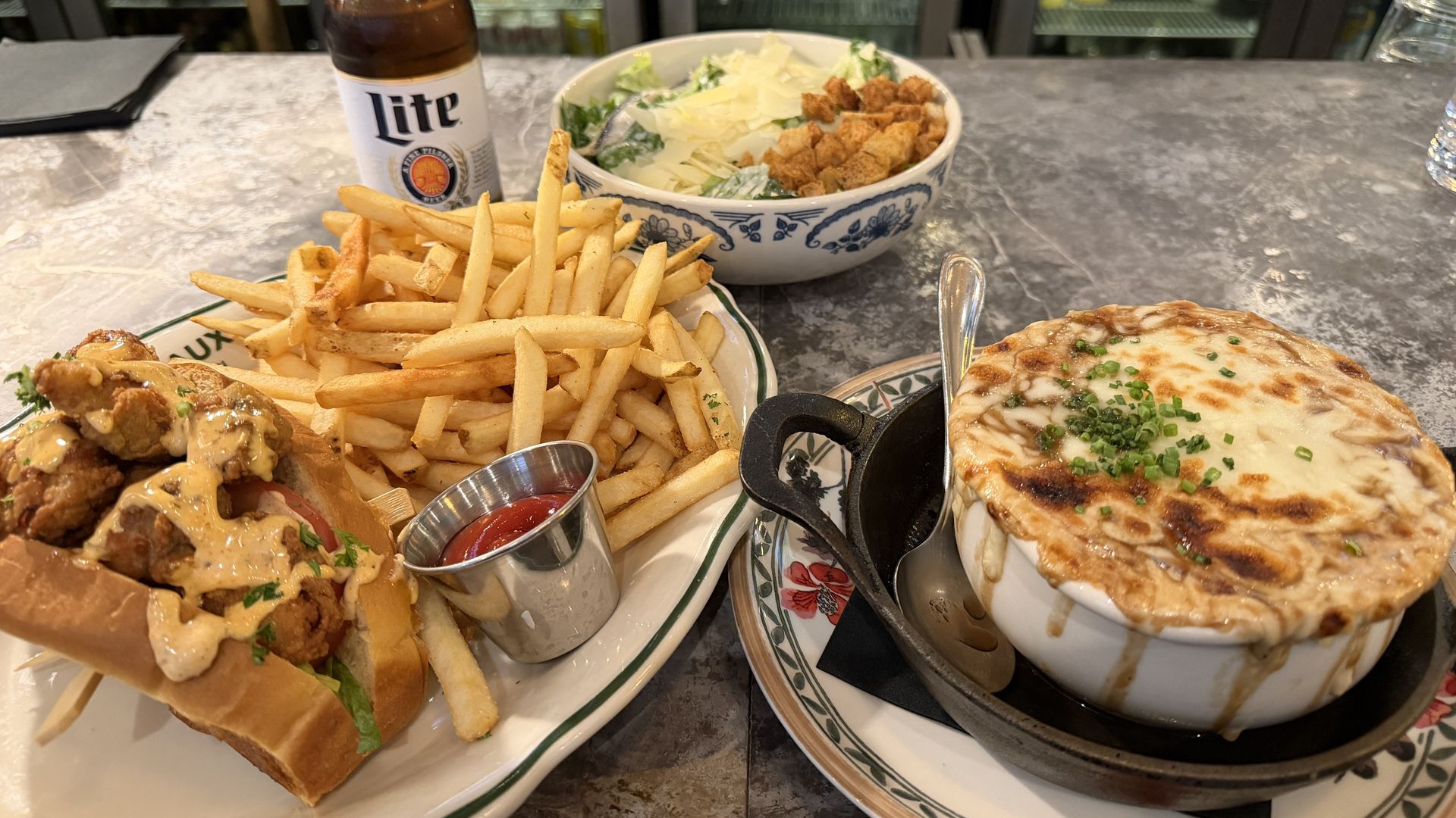 Bar counter meal: Miller Lite bottle, plate of fries with a fried chicken sandwich and creamy sauce, a side salad, and baked macaroni and cheese with chives in a ramekin.