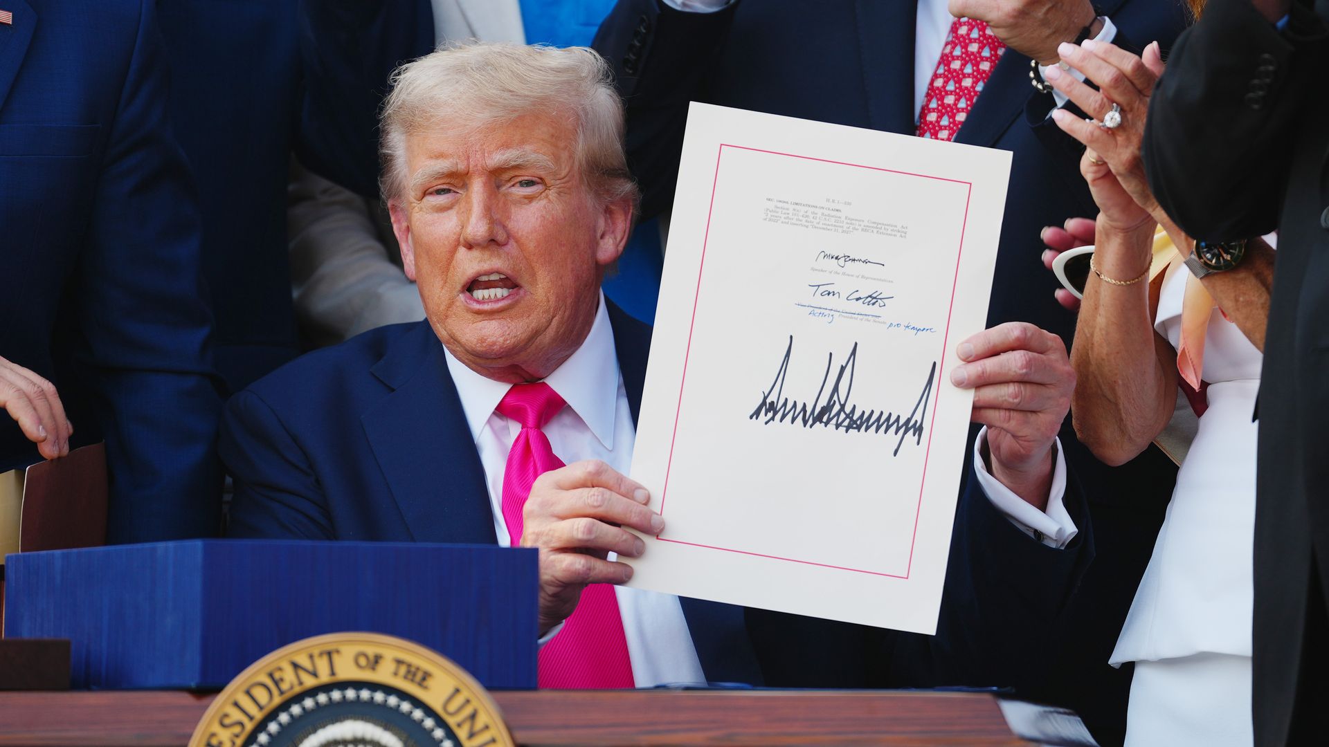 Former President Donald Trump in a dark suit and bright pink tie holding a signed document, surrounded by applauding people in business attire, with a presidential seal visible on the desk.