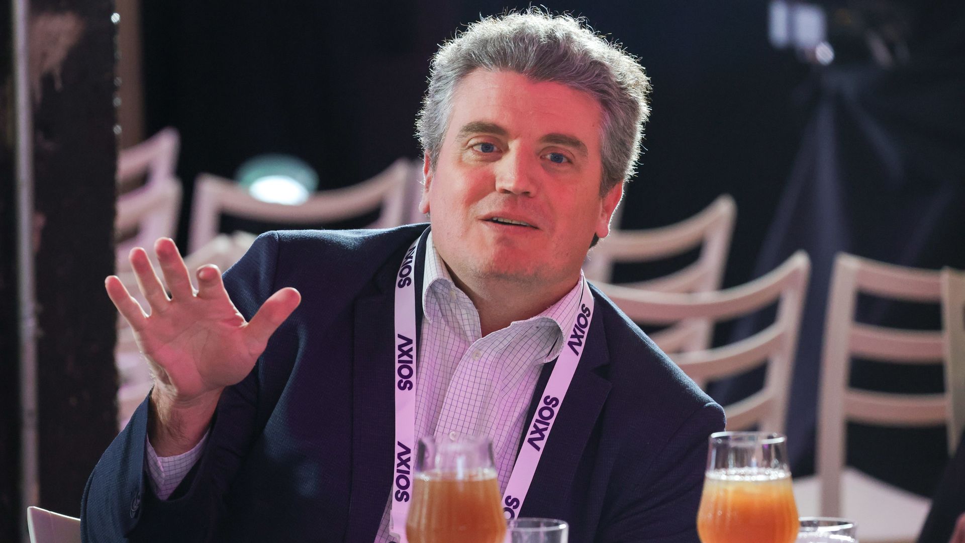 Man in a dark suit sits at a table, gesturing with his left hand. He wears a lanyard; two orange drinks sit in front, with chairs in the background.