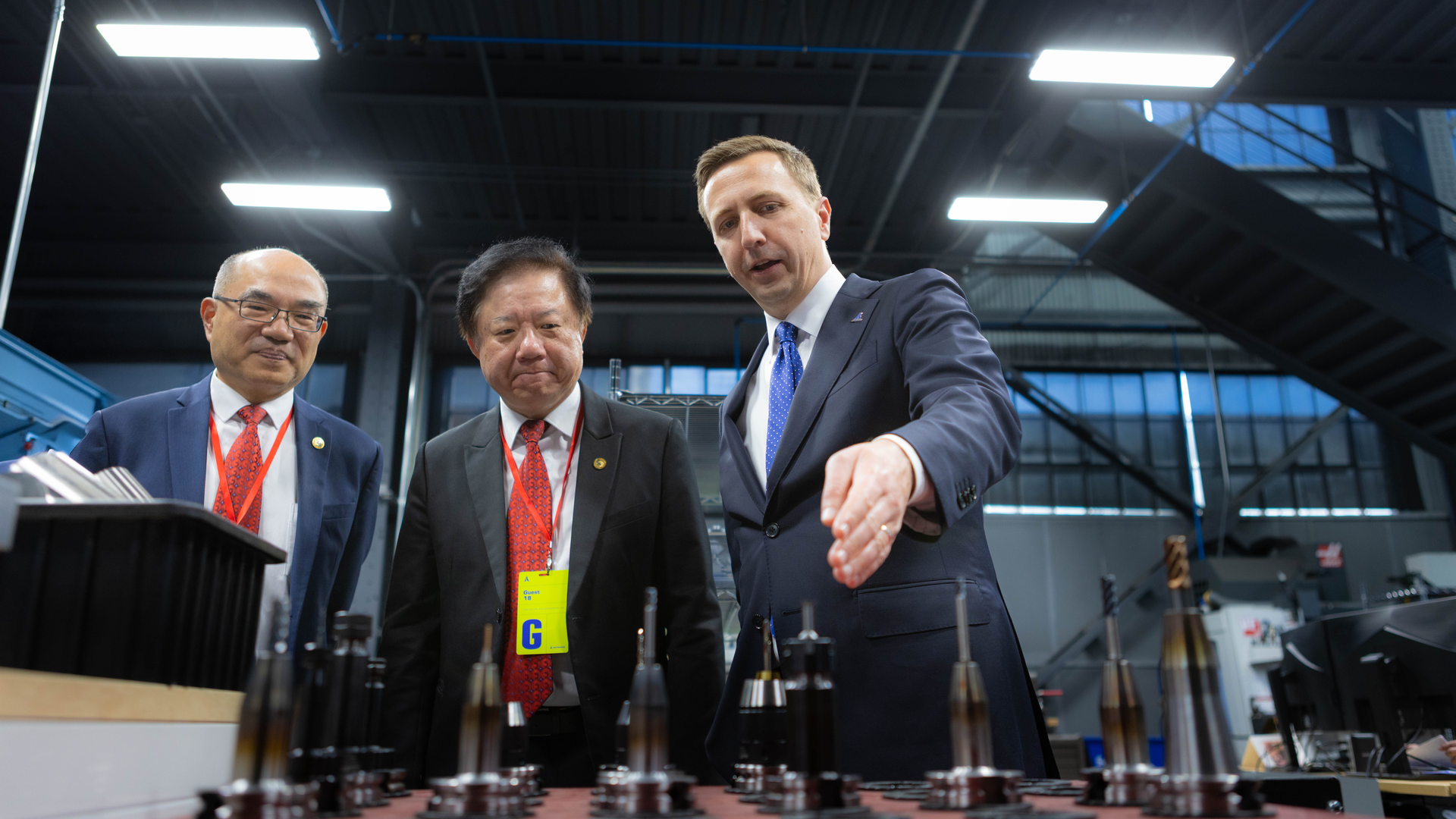 Three men stand in a renovated warehouse. They are all wearing suits with ties. The man on the far right is gesturing toward a table in the foreground.
