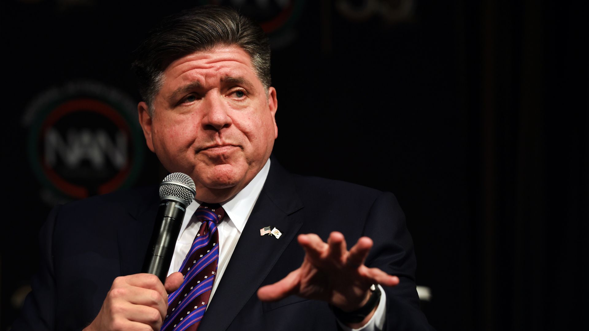 A man in a dark suit and striped tie holds a microphone on a dark stage, gesturing with his hands; small American flag lapel pins are visible.