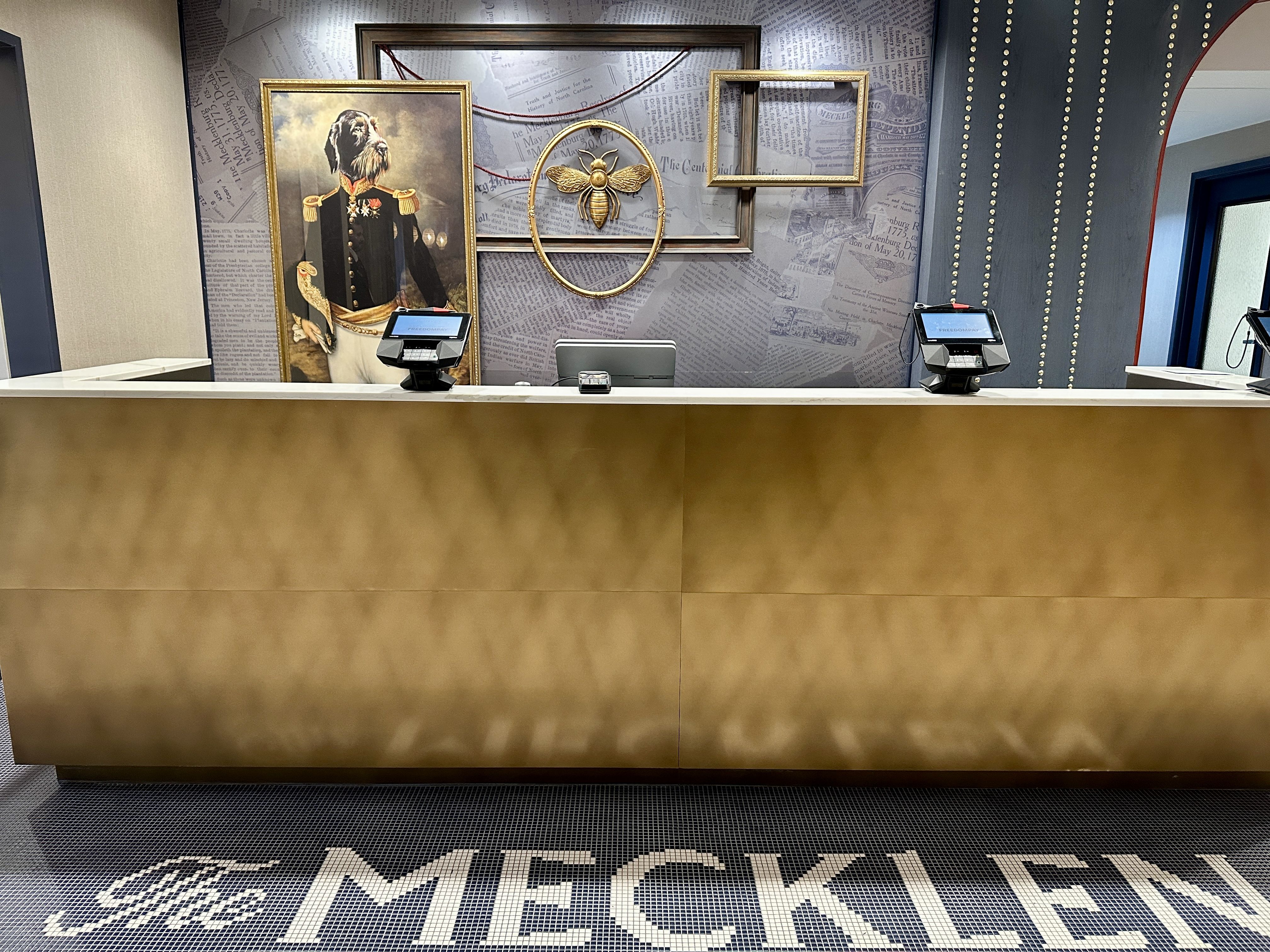 Reception desk with gold front, two computer screens, a framed painting of a dog in a military uniform, empty picture frames, and a large gold bee decoration on a newspaper-textured wall. "The MECKLEN" floor mosaic.