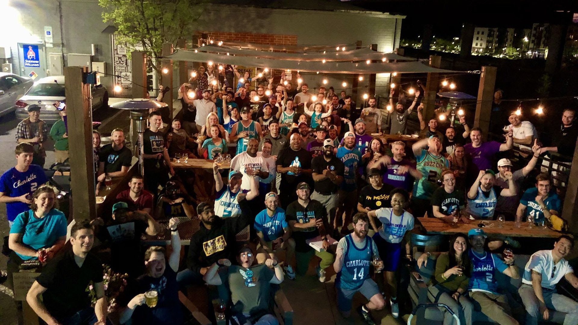 Large group of people at a nighttime rooftop party, many wearing teal Charlotte jerseys, posing and cheering under string lights with drinks, wooden tables, and an urban backdrop.