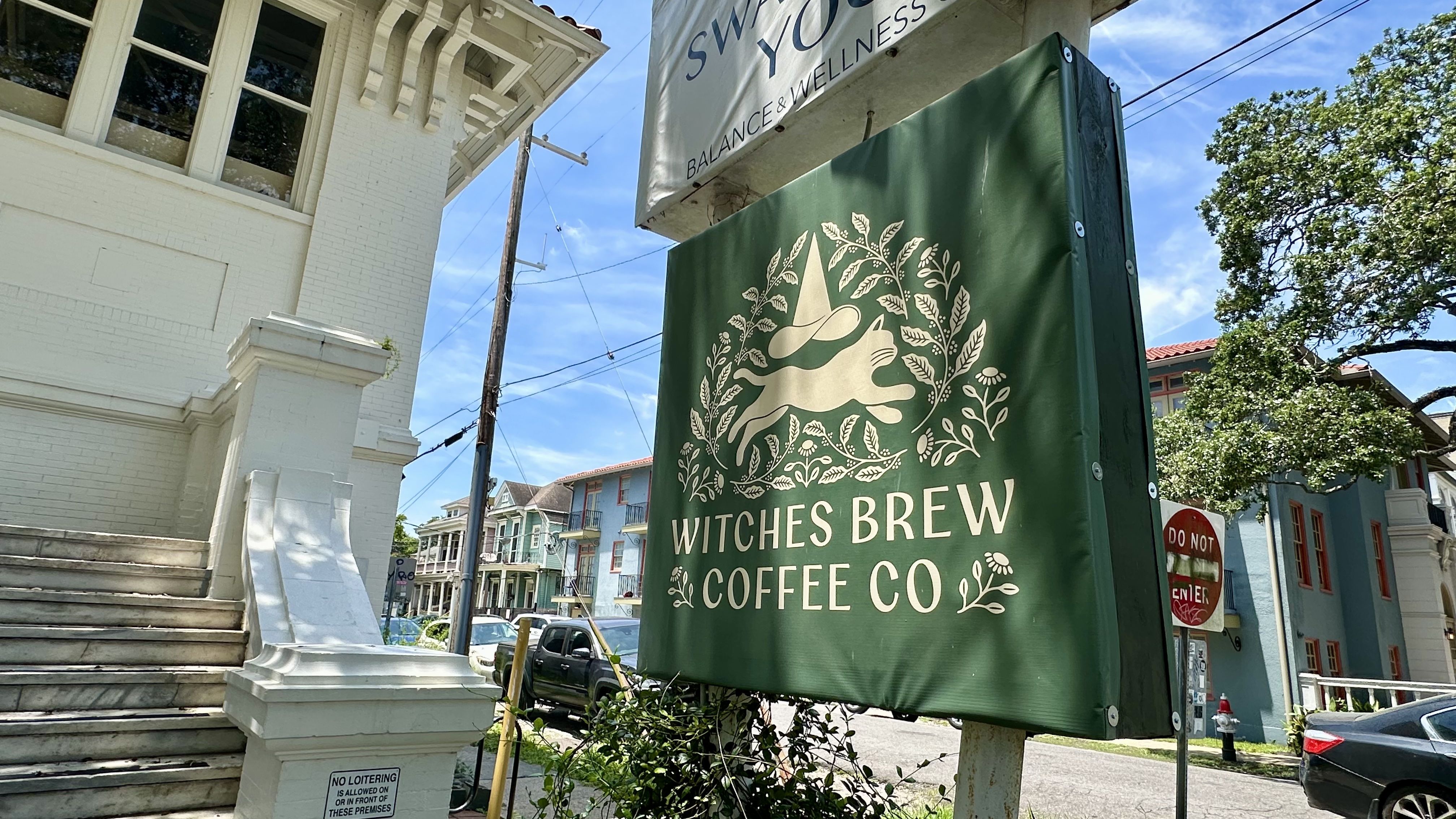 White brick building with a decorative cornice on the left, a large green banner reading WITCHES BREW COFFEE CO with a white illustrated witch and leaves, against a blue sky and trees.