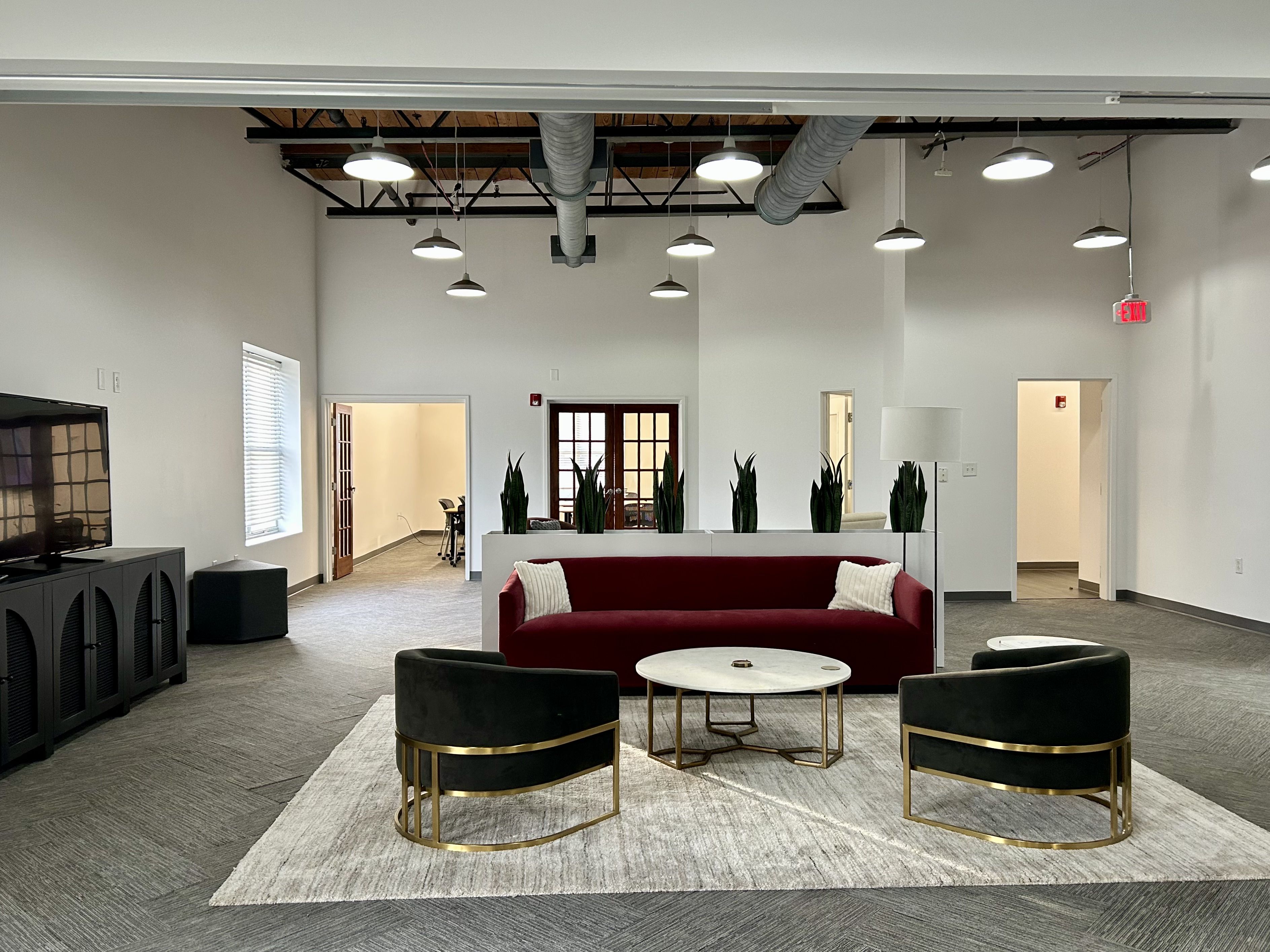 Modern office lounge with a burgundy sofa, two black and gold chairs, a round white marble coffee table on a gray rug, white walls, hanging lights, and potted plants dividing the space.