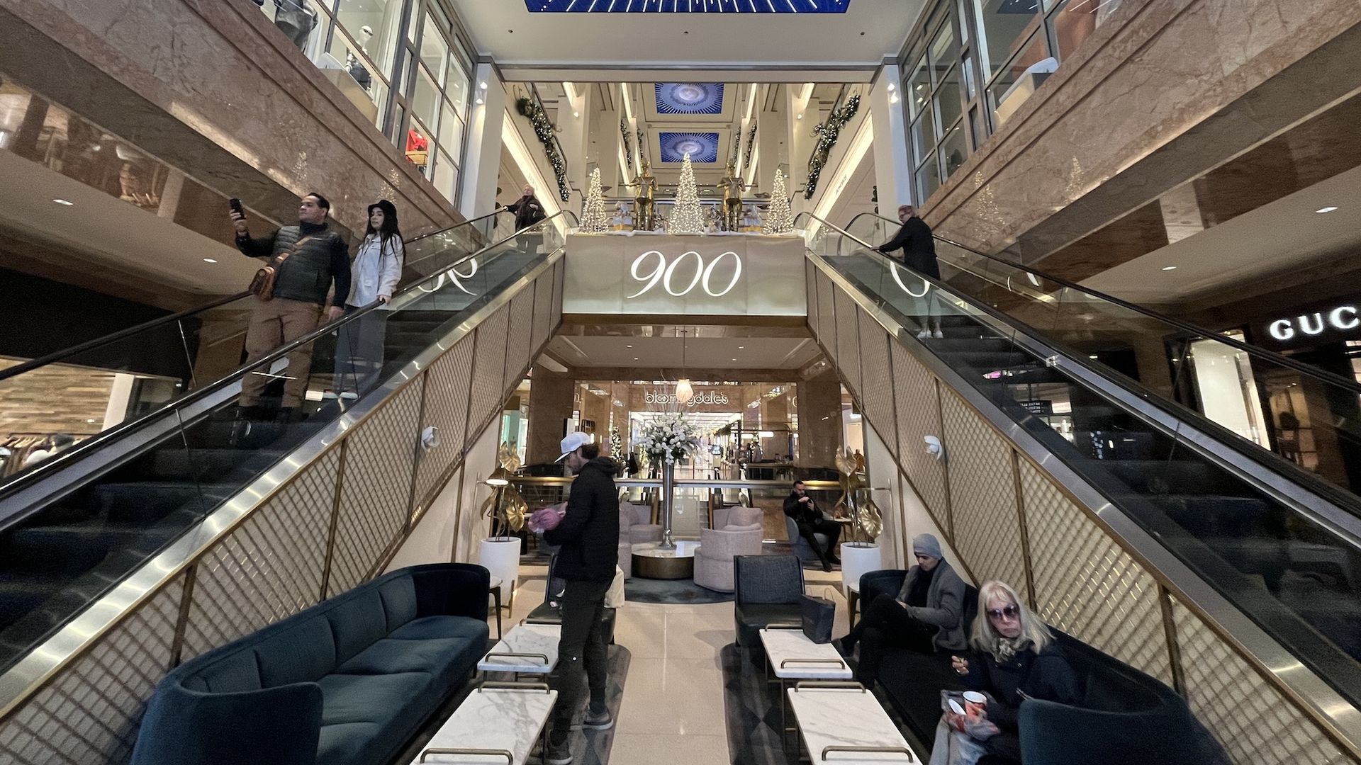 Two escalators on the sides of a gold sign reading 900 with Bloomingdale's in the background and lit Christmas tree.