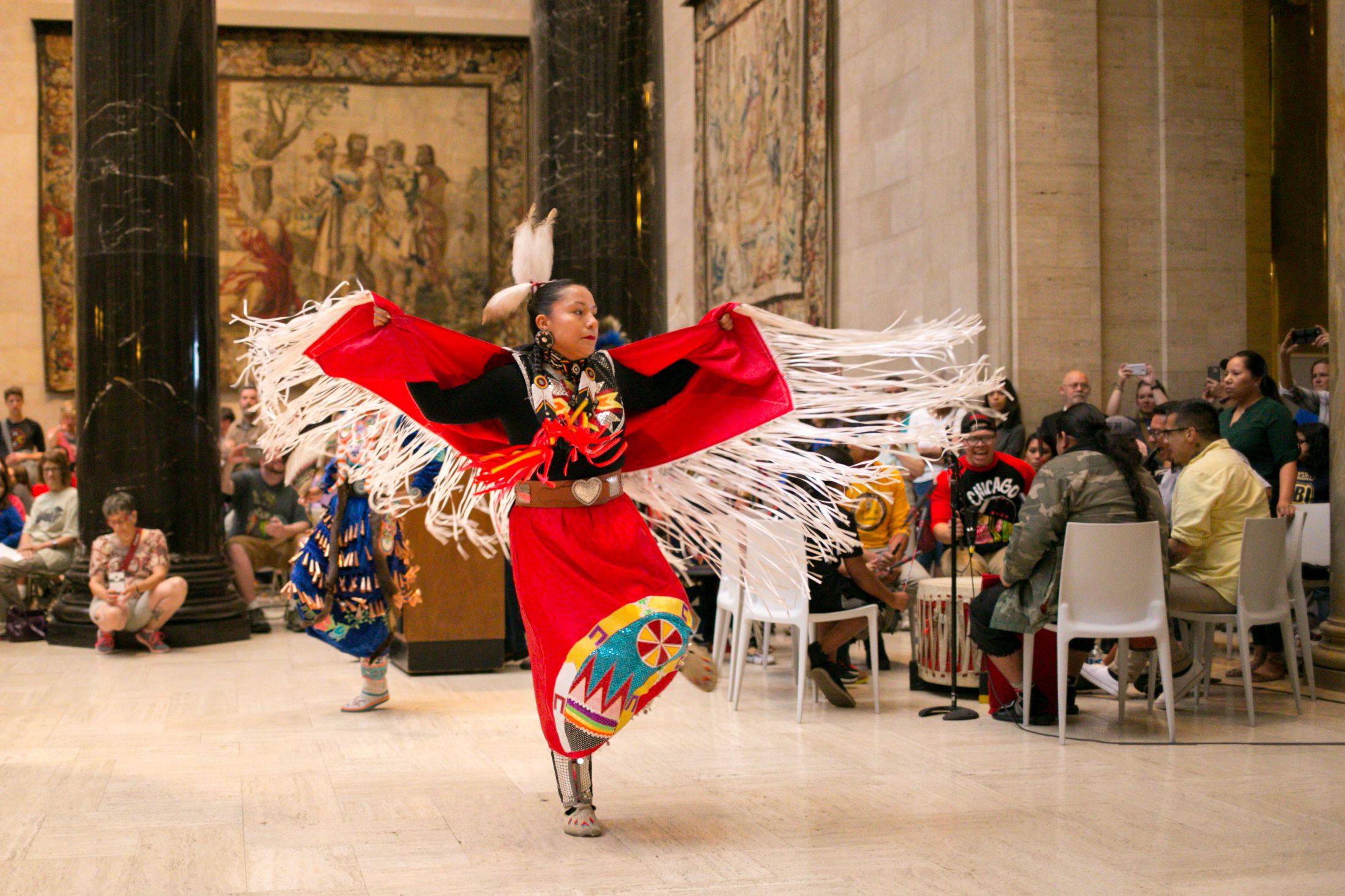 A dancer in red traditional Native American attire with white fringe and beadwork performs indoors, surrounded by an audience and musicians playing drums in a room with large black columns and tapestries.