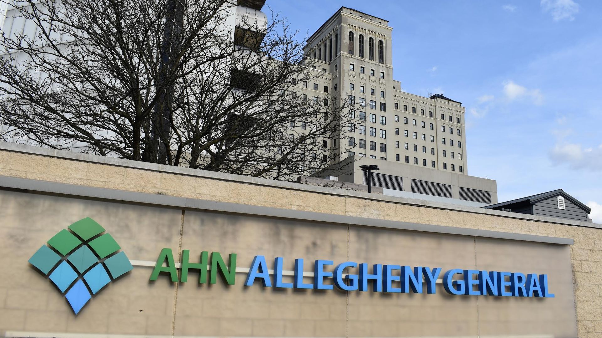 Outdoor view of AHN Allegheny General hospital sign with green-blue diamond logo on a beige wall, a tall beige building in the background, and a leafless tree against a bright blue sky.