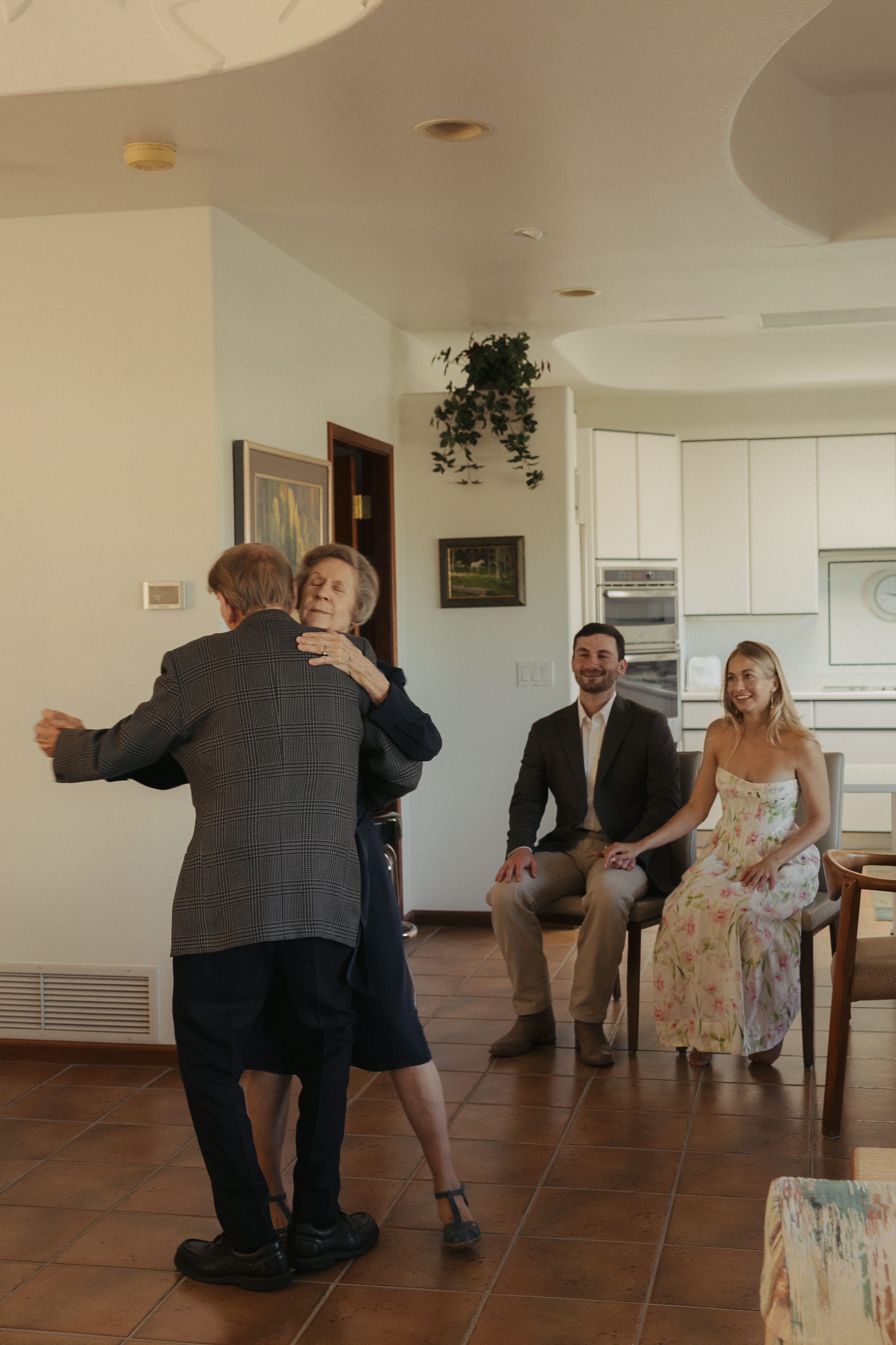 Two older adults dancing in a bright kitchen area while a man in a suit and a smiling woman in a floral dress sit nearby; white cabinets and wall art are visible.