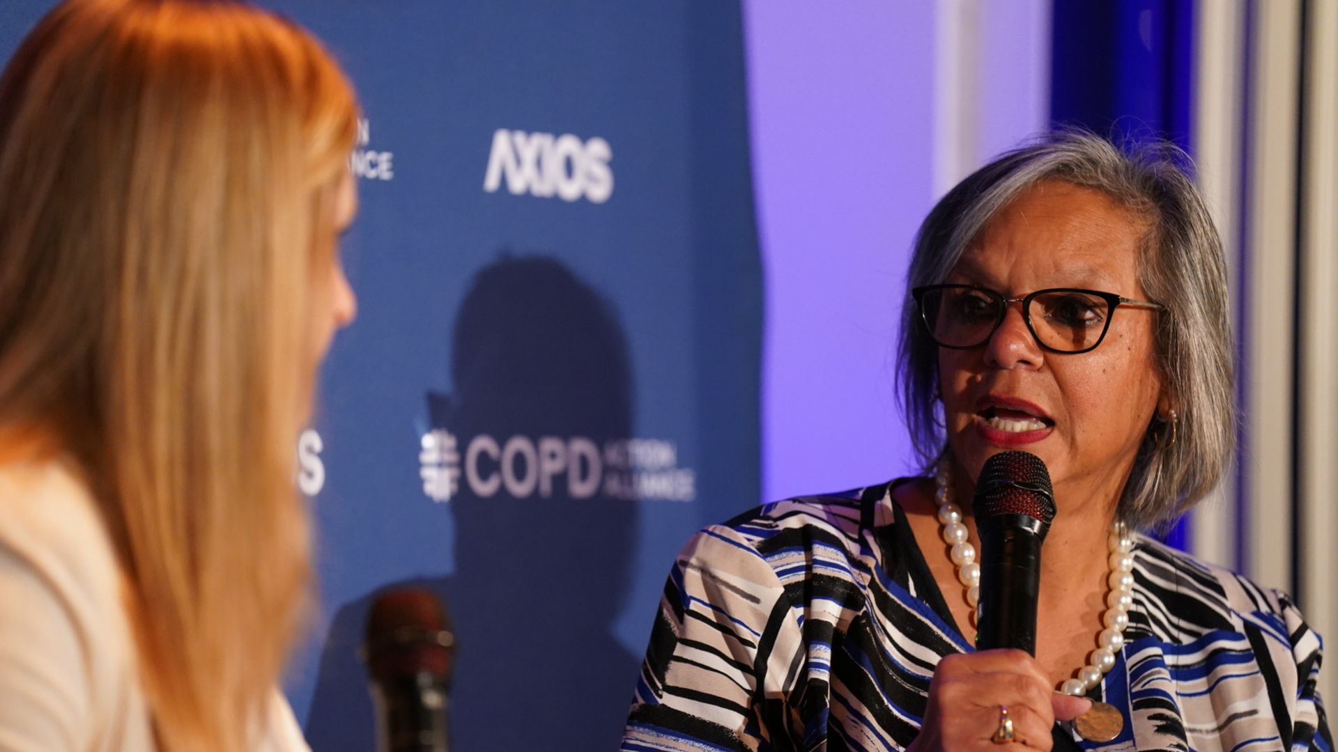 Two women holding microphones engaged in conversation on stage; one has gray hair and glasses wearing a blue, white, and black patterned jacket with a pearl necklace; blue backdrop with "AXIOS" and "COPD" logos.