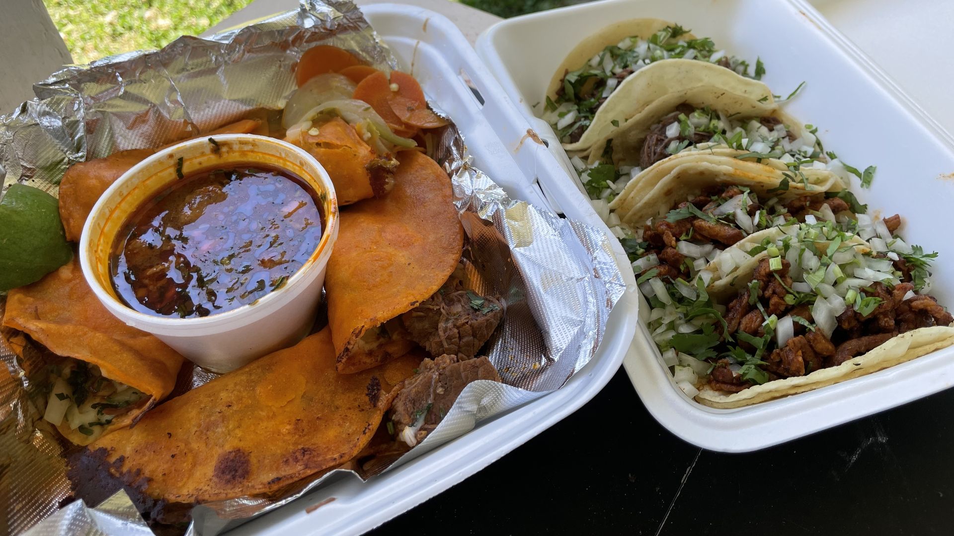 Two takeout trays outdoors: left shows orange tortillas with a cup of red sauce and foil, right holds tacos topped with onions and cilantro, on a grassy background.