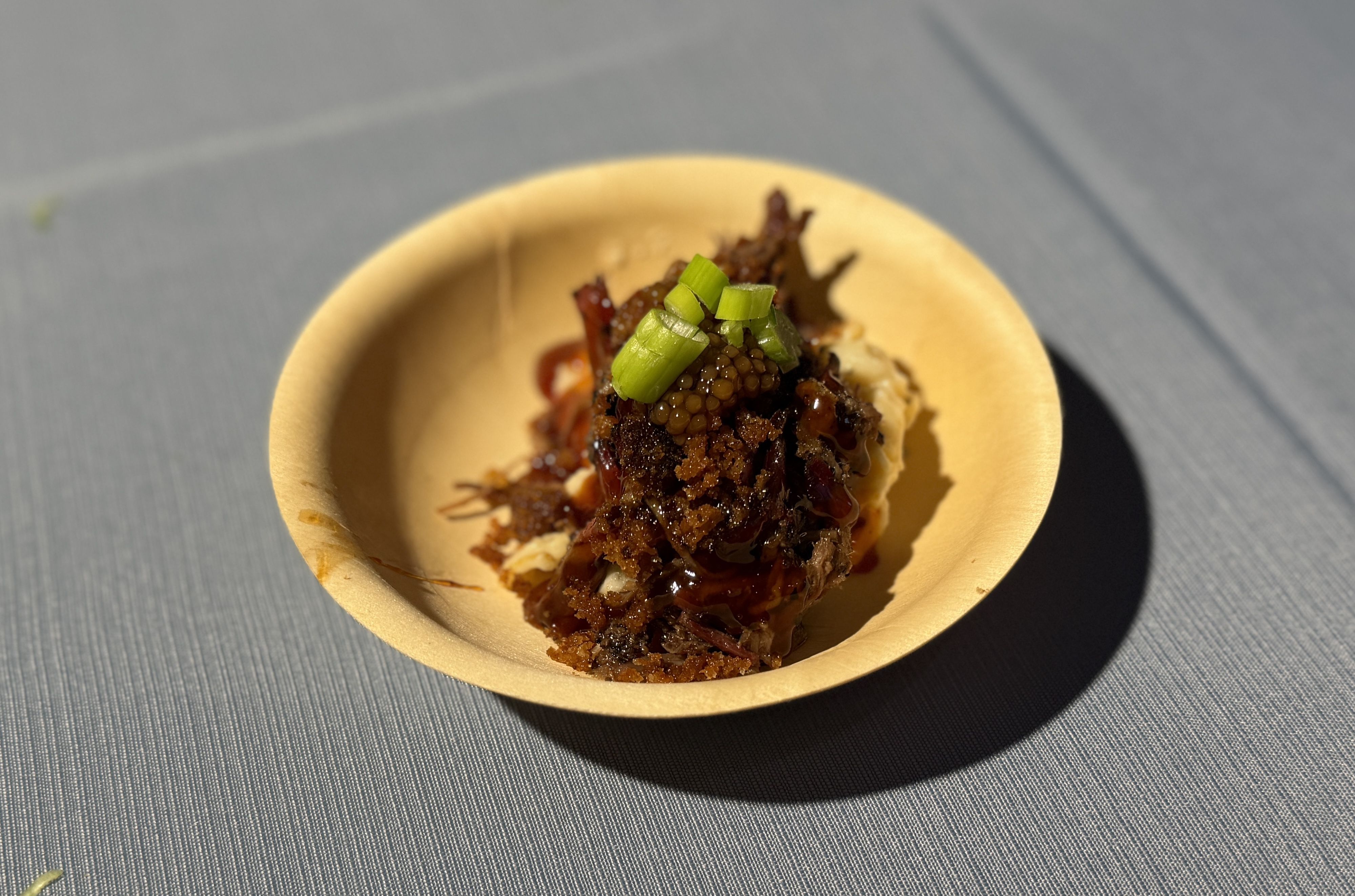 Small beige paper cup holds a mound of caramelized, glossy meat with crunchy bits, topped with chopped green onions, set on a blue-gray textured surface.