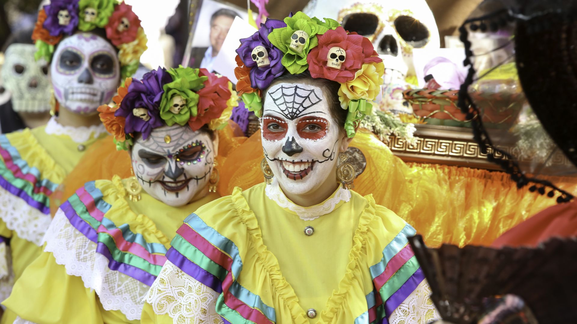 Three young women, with their faces painted as Mexican sugar skulls, stand in a line wearing ballet folklorico dresses. 