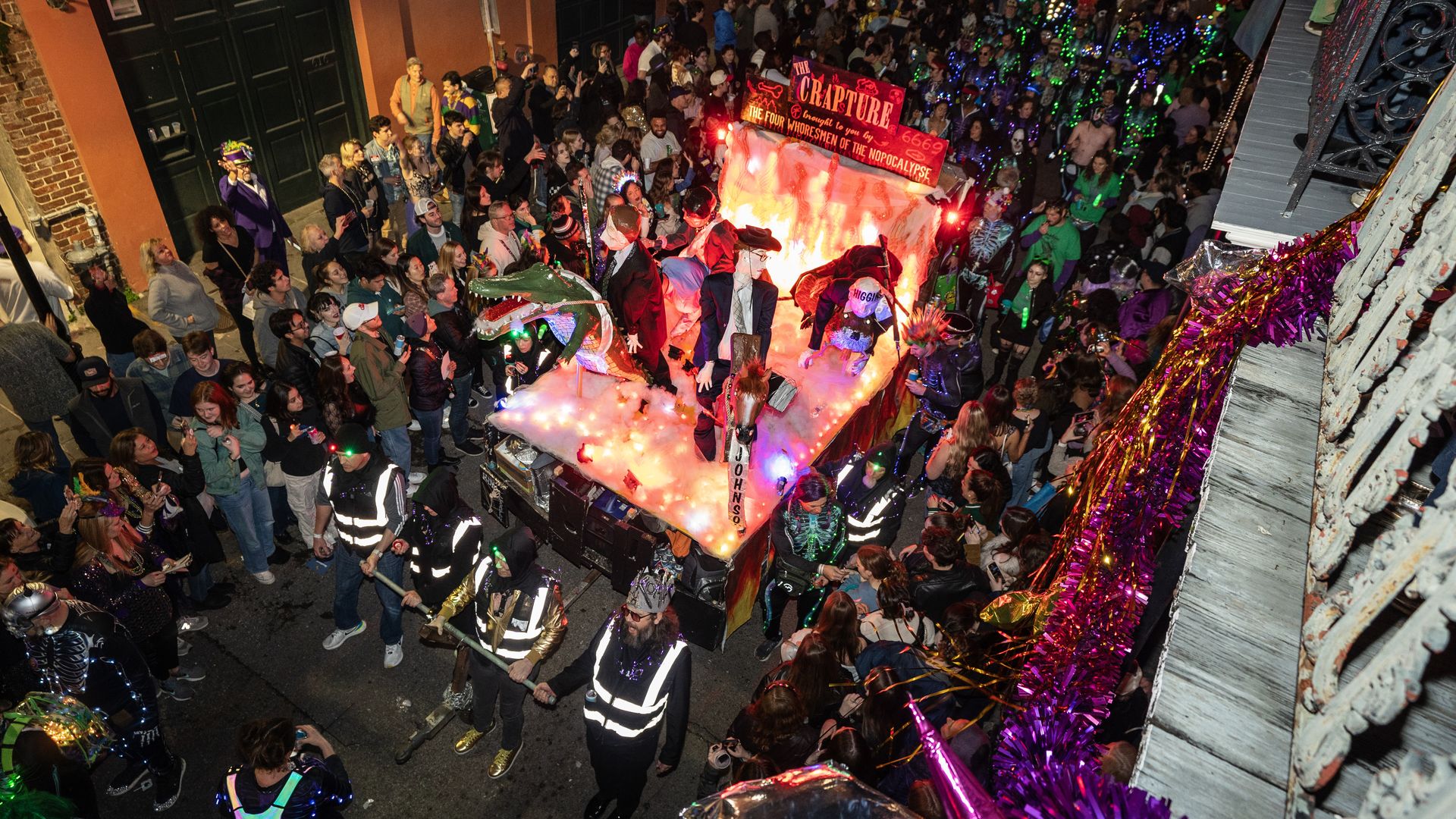 A Krewe du Vieux float is viewed from a balcony above the crowded parade route.