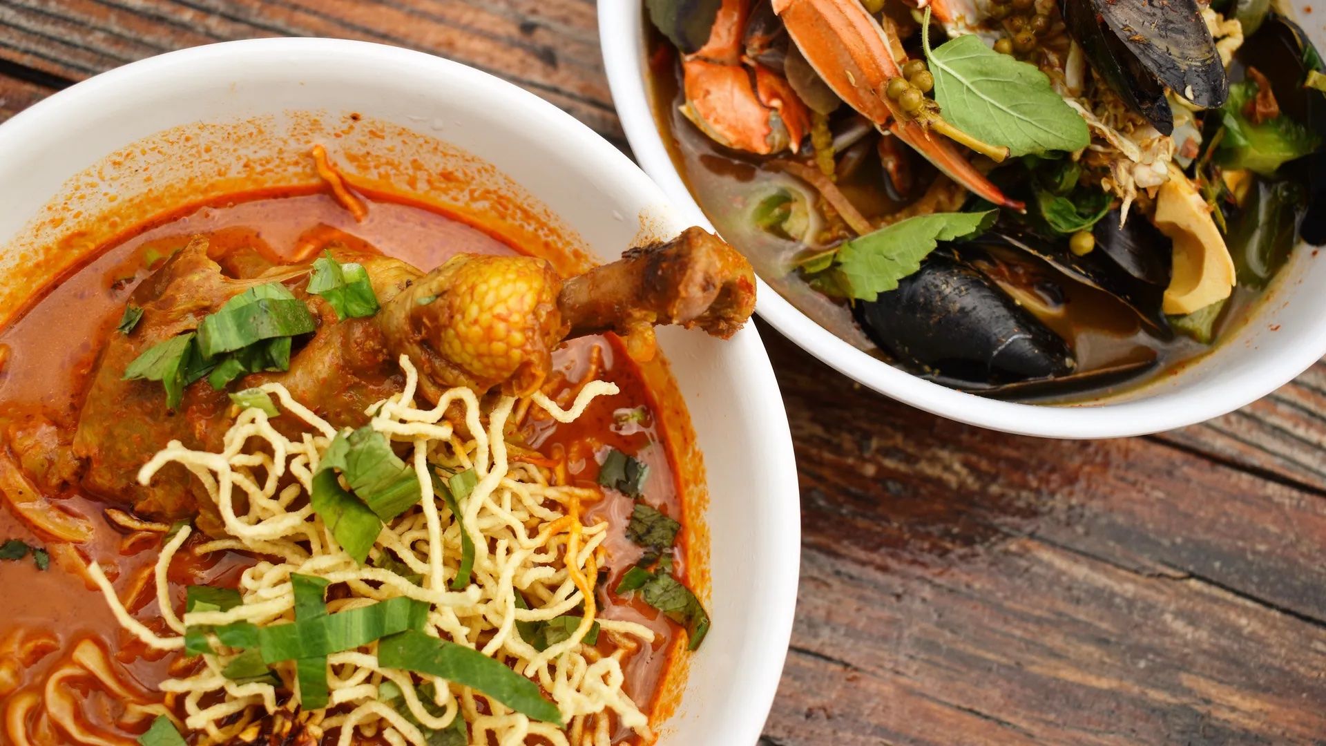 Two bowls of soup on a wooden surface; one contains red curry with chicken drumstick, crispy noodles, and green herbs, the other has seafood including crab, mussels, and green leaves.