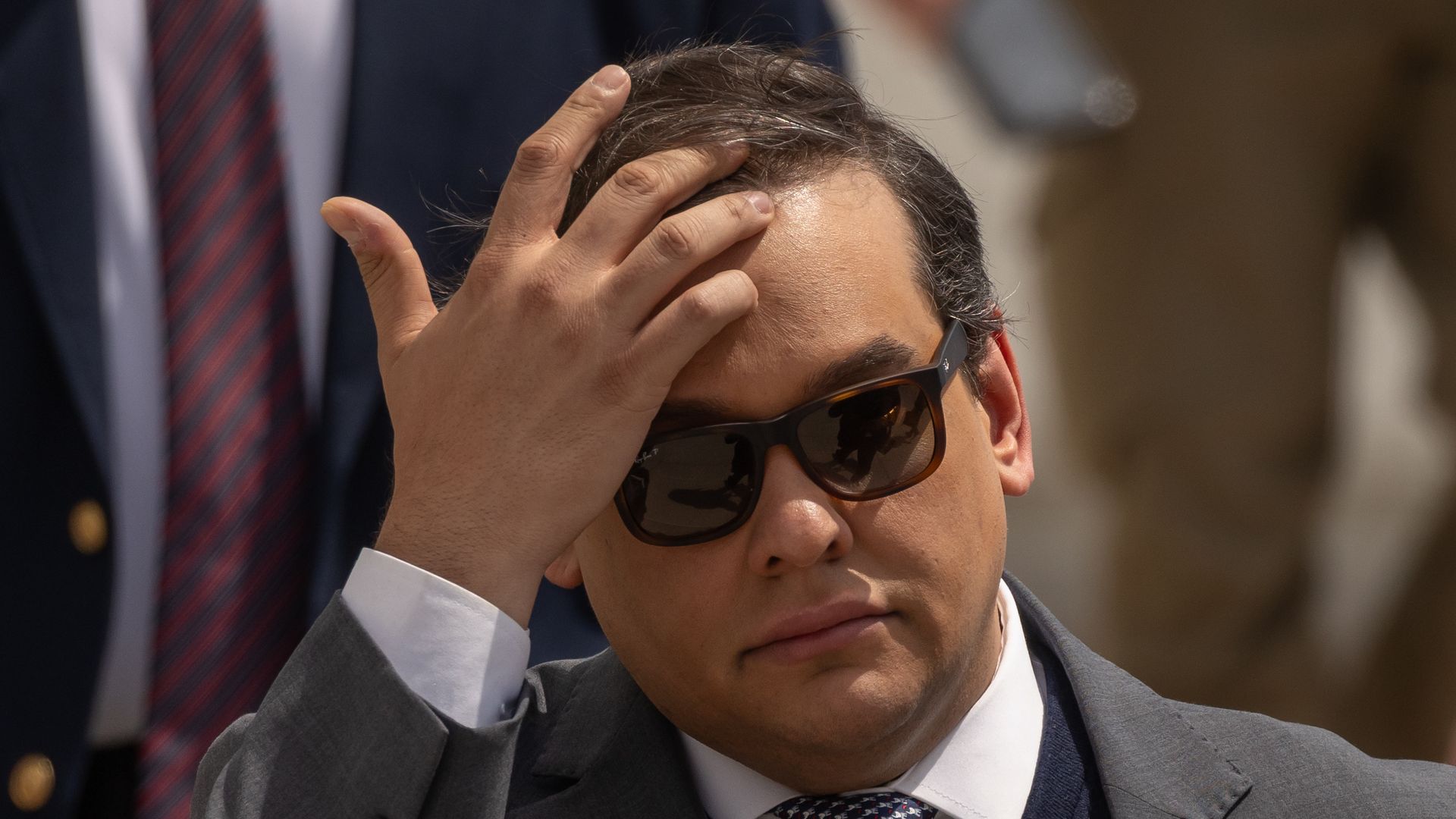 George Santos wears sunglasses and a grey jacket, while touching his hair. 