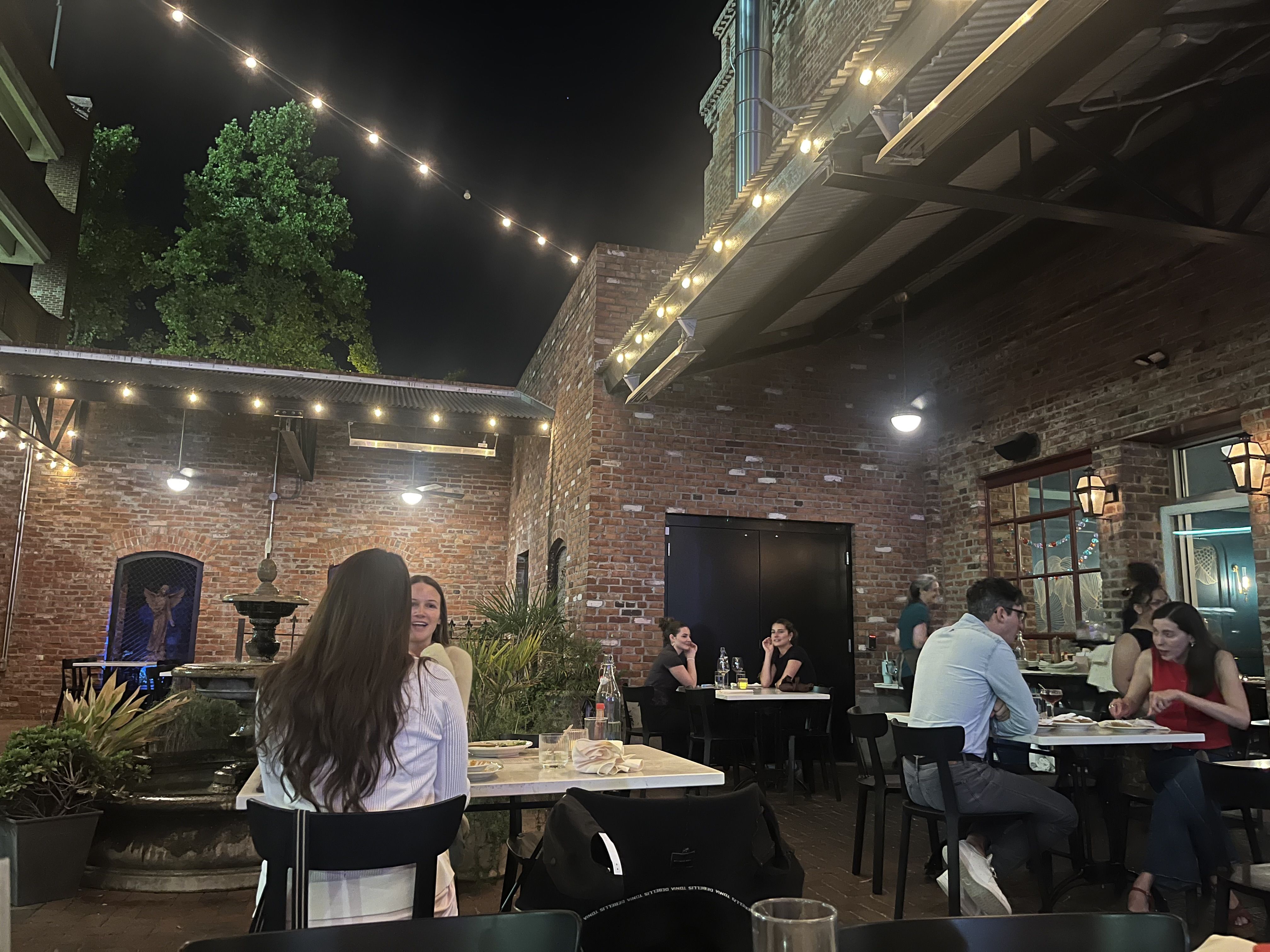 Nighttime outdoor restaurant courtyard with brick walls, string lights overhead, and groups of diners at tables. A green tree on the left and a fountain in the foreground.