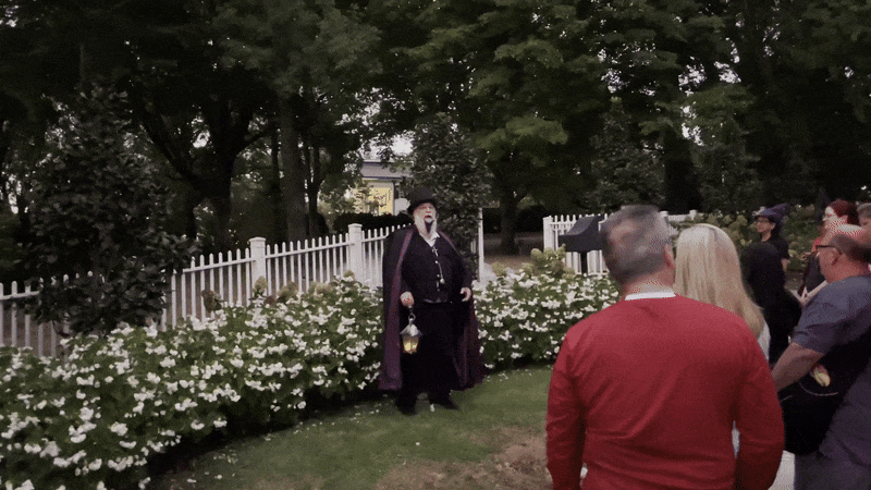 A tour guide dressed in a Halloween outfit turns and points into the woods during a tour while a small crowd looks on. 