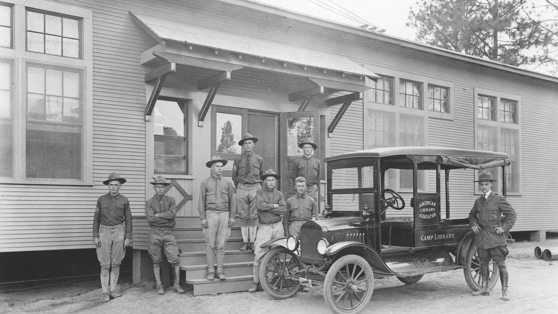 A black and white photo of soldiers posing on the steps in front of a building, with an early-model car parked out front.