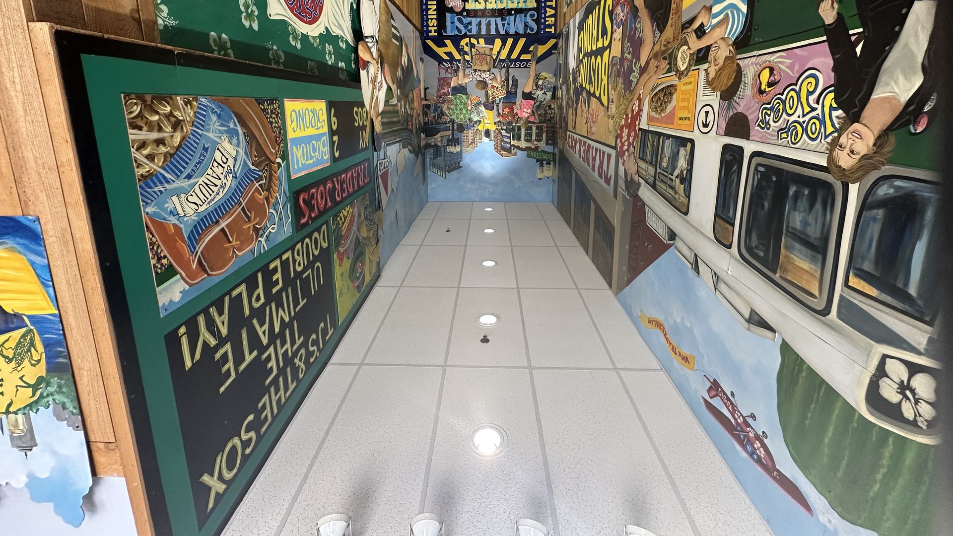 Colorful mural in a hallway depicting Boston sports, a bus, people, and signs with vibrant text and images on wooden walls and ceiling lights above.