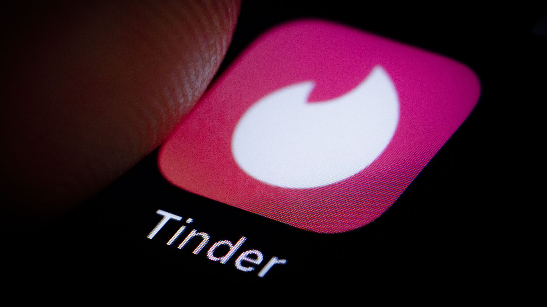 In this image, a finger hovers over the Tinder app logo on a persons phone.
