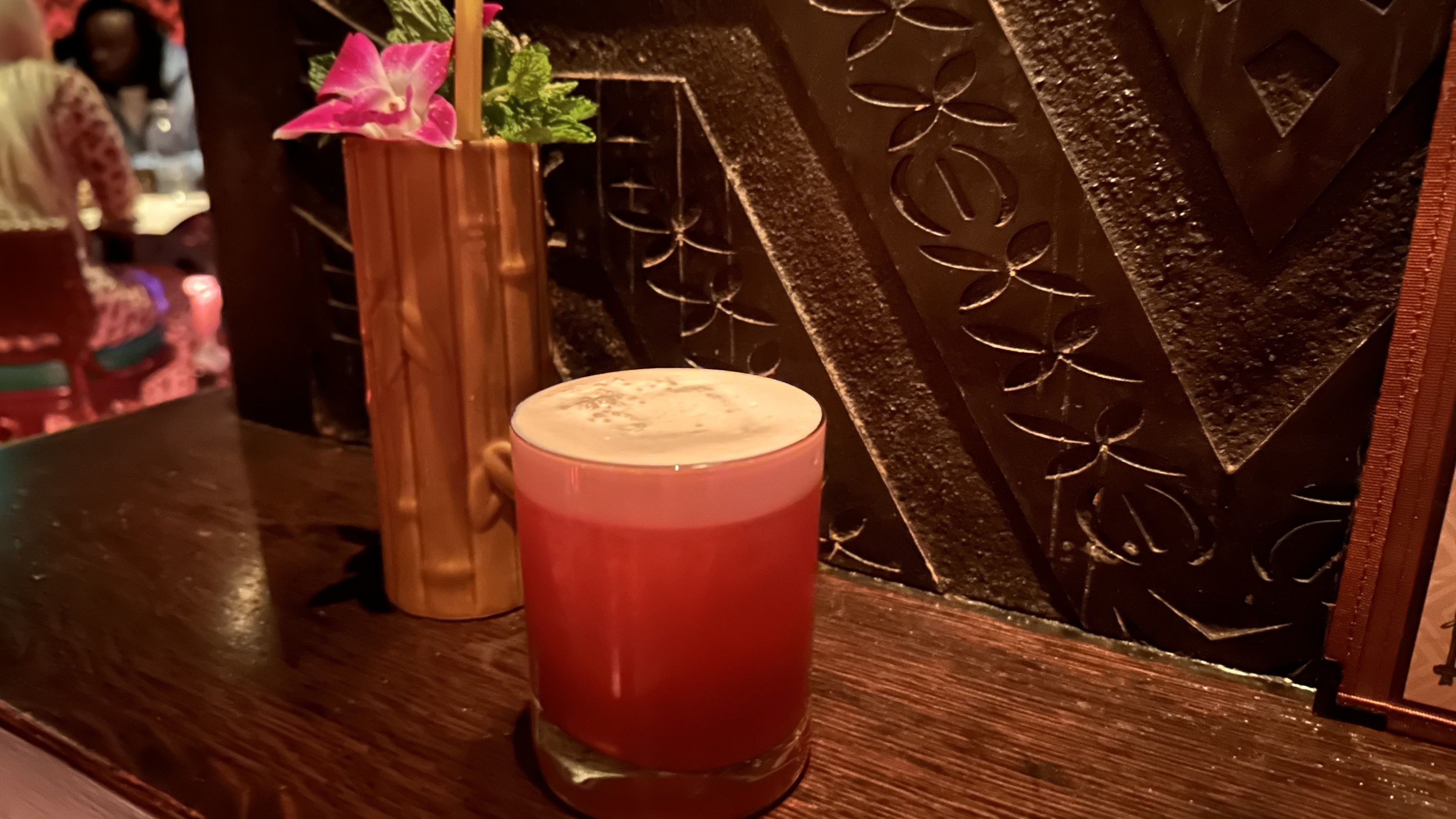 Dimly lit bar scene featuring a red layered cocktail with foam on a wooden table. A cocktail in a bamboo glass topped with a pink flower with pink flowers sits beside; a dark carved wall and blurred patrons fill the background.
