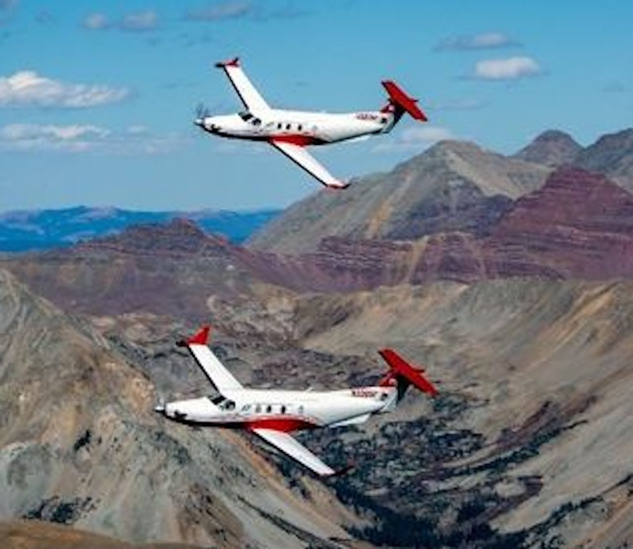 Two white private jets with red tails fly in formation over rugged brown-gray canyon mountains beneath a clear blue sky.