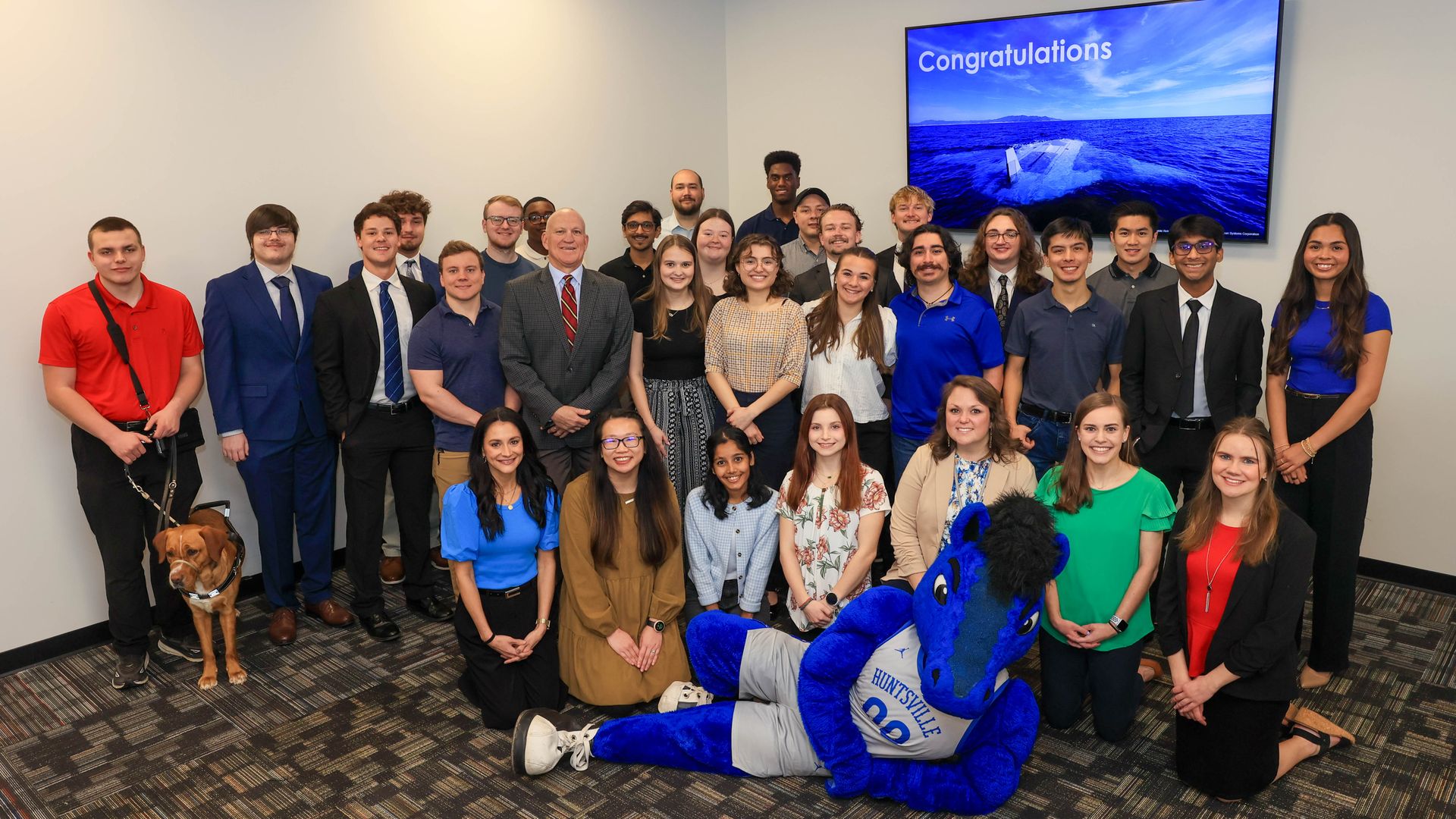 Group of diverse students and a suited presenter pose in a room; a blue mascot lies on the carpet, a leashed brown dog stands left, and a screen reads "Congratulations" over a sea image.