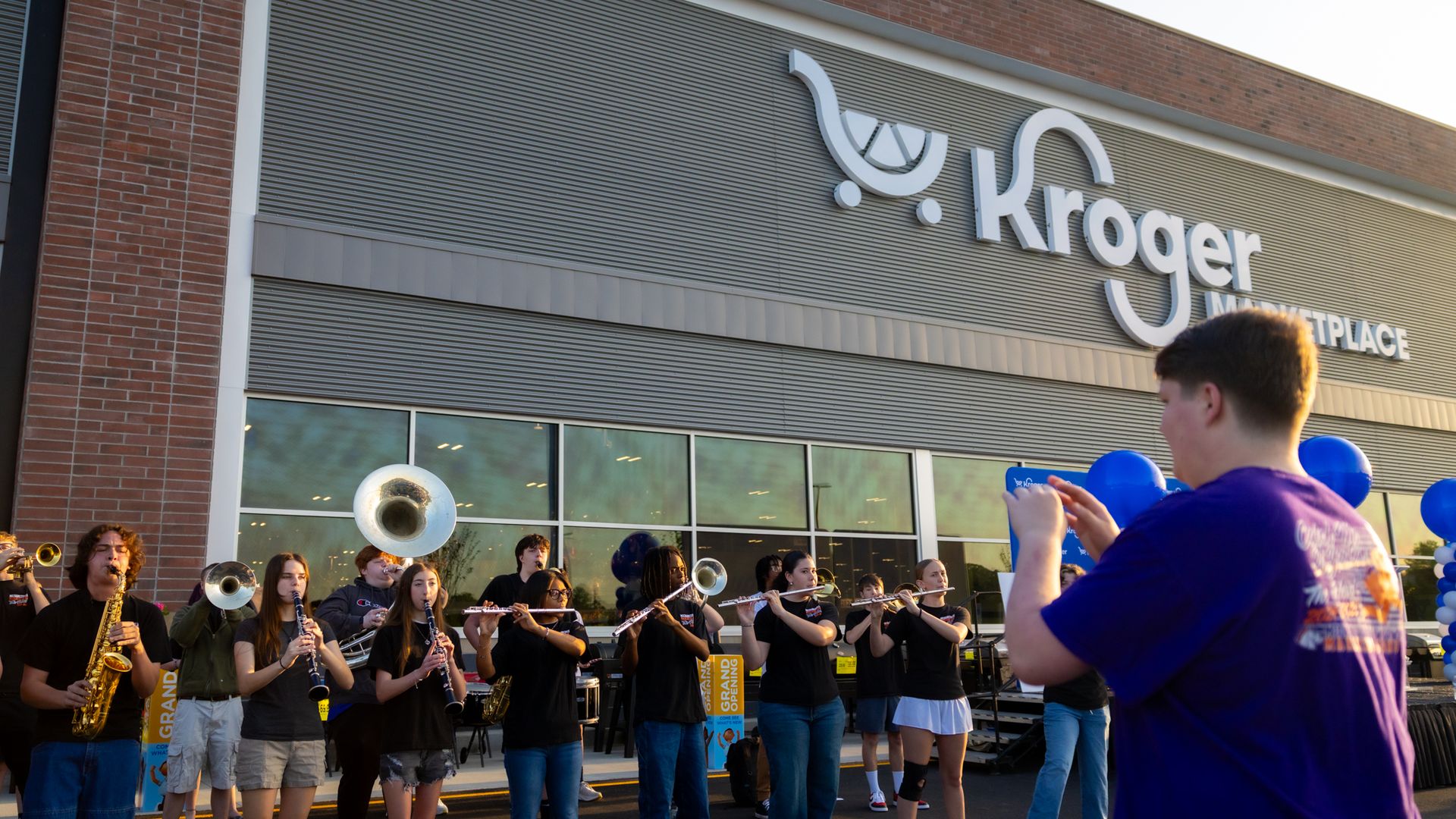 A student band performs outside Kroger Marketplace, playing brass and woodwind instruments as a person in a purple shirt records the scene.