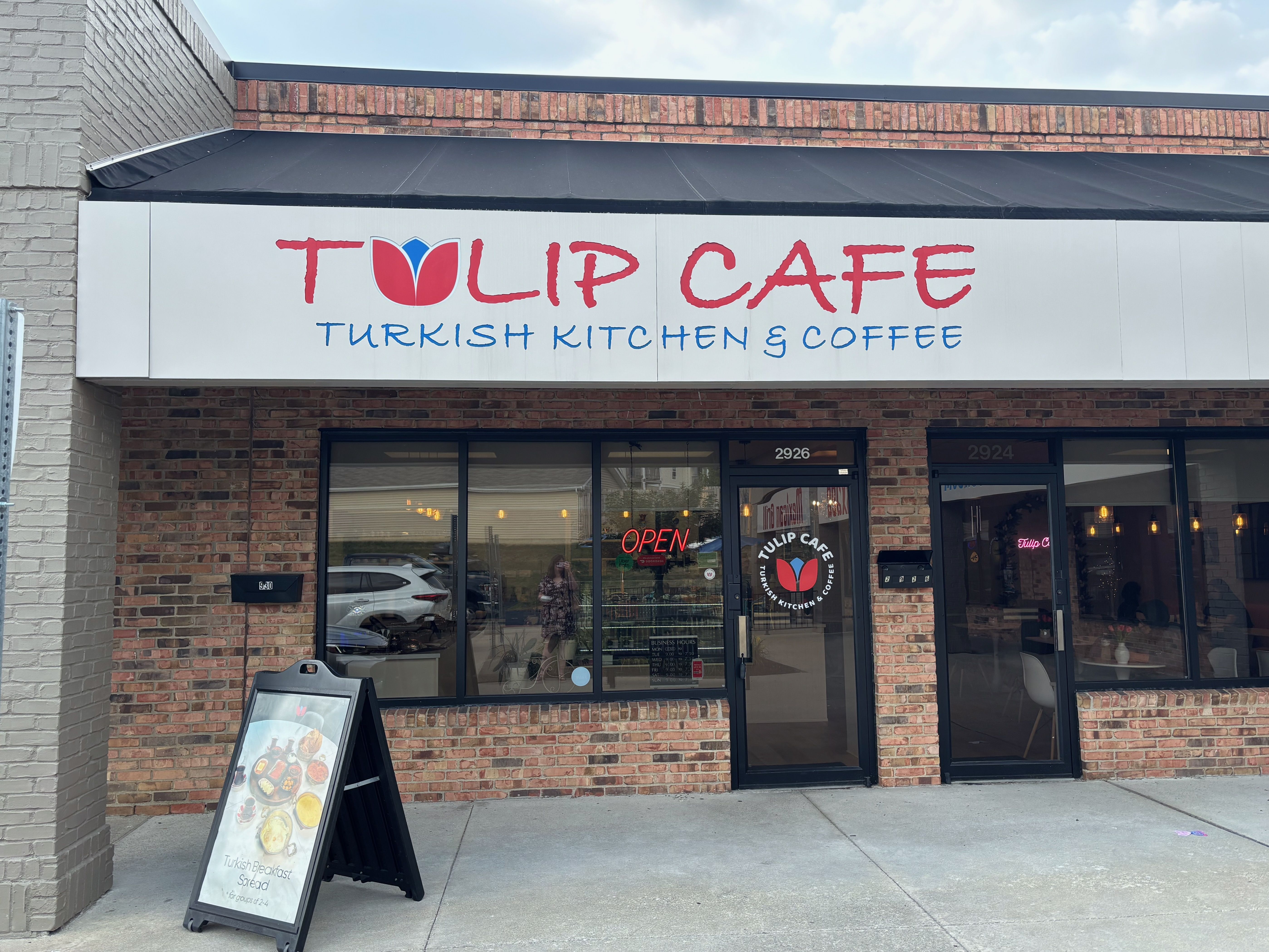Front of Tulip Cafe storefront: white sign with red Tulip Cafe lettering and a red tulip logo with blue center; blue Turkish Kitchen & Coffee text beneath; brick facade, black awning, glass doors, OPEN sign.