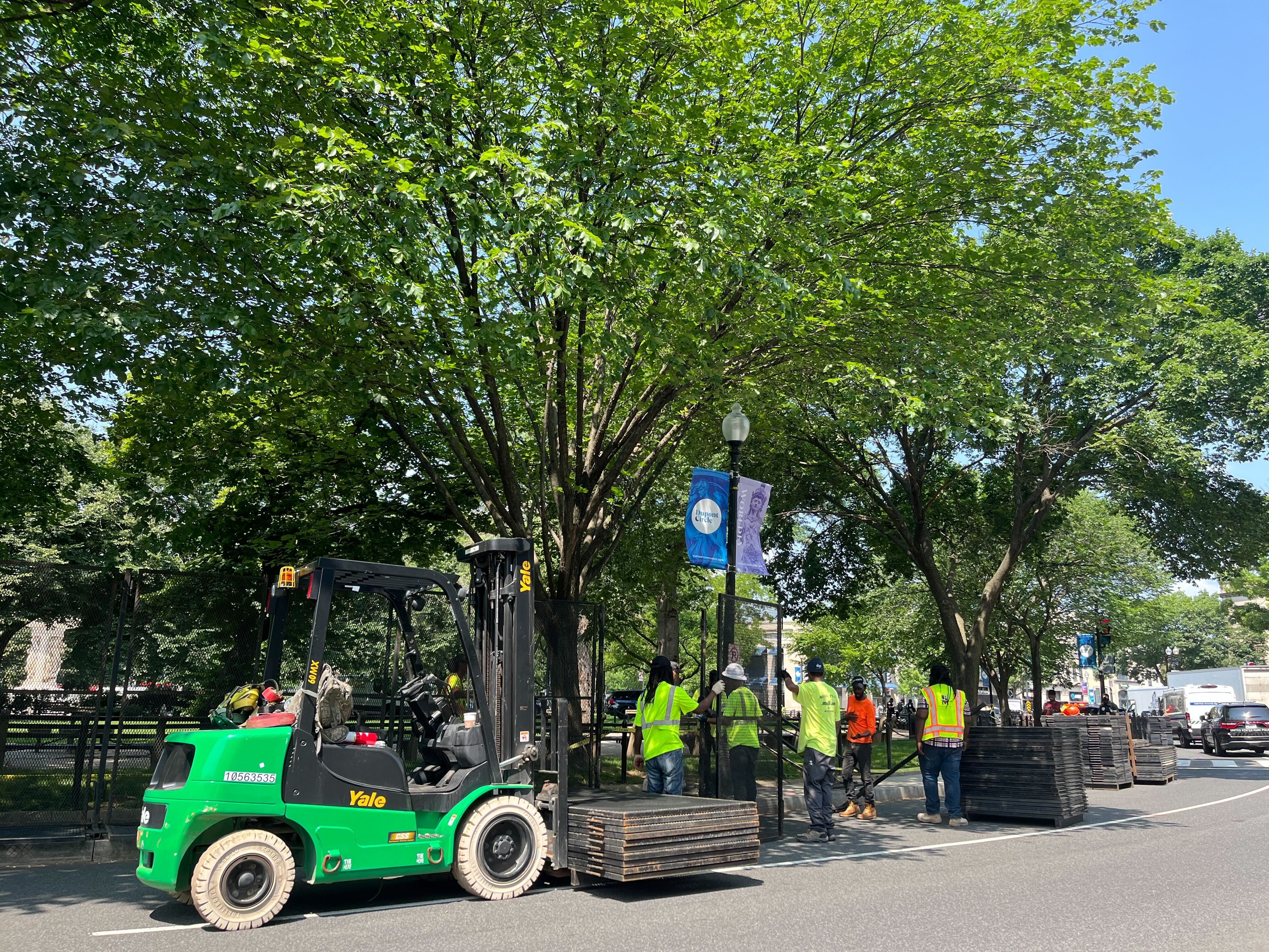 A green service vehicle and work crews in hardhats and flourescent shirts install black fencing around the park. 