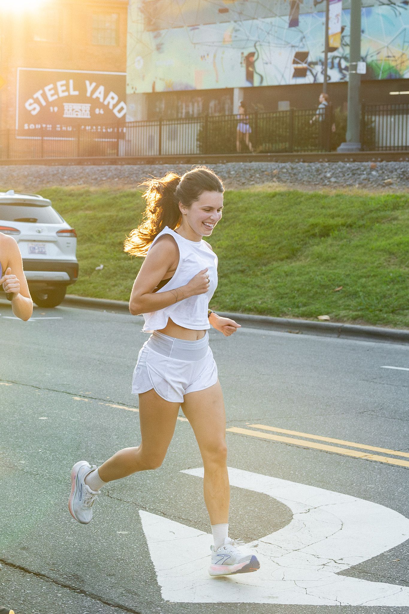 A woman in white athletic wear runs on a sunlit city street; behind her is a colorful mural, a barrier fence, the sign "STEEL YARD", and a parked car.