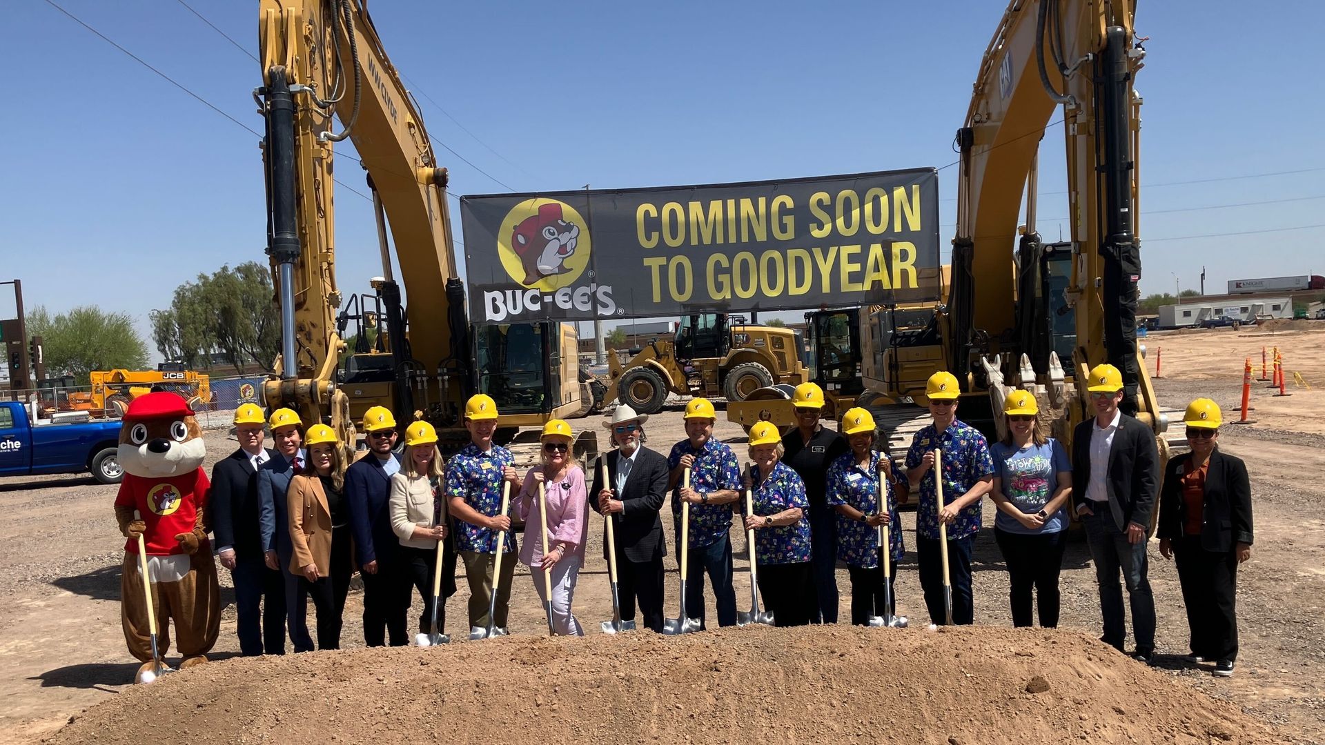 Seventeen people stand in a row holding shovels in front of a mound of dirt, the furthest-most left one wearing a beaver costume. A banner announcing an incoming Buc-ee's is between two backhoes behind them.