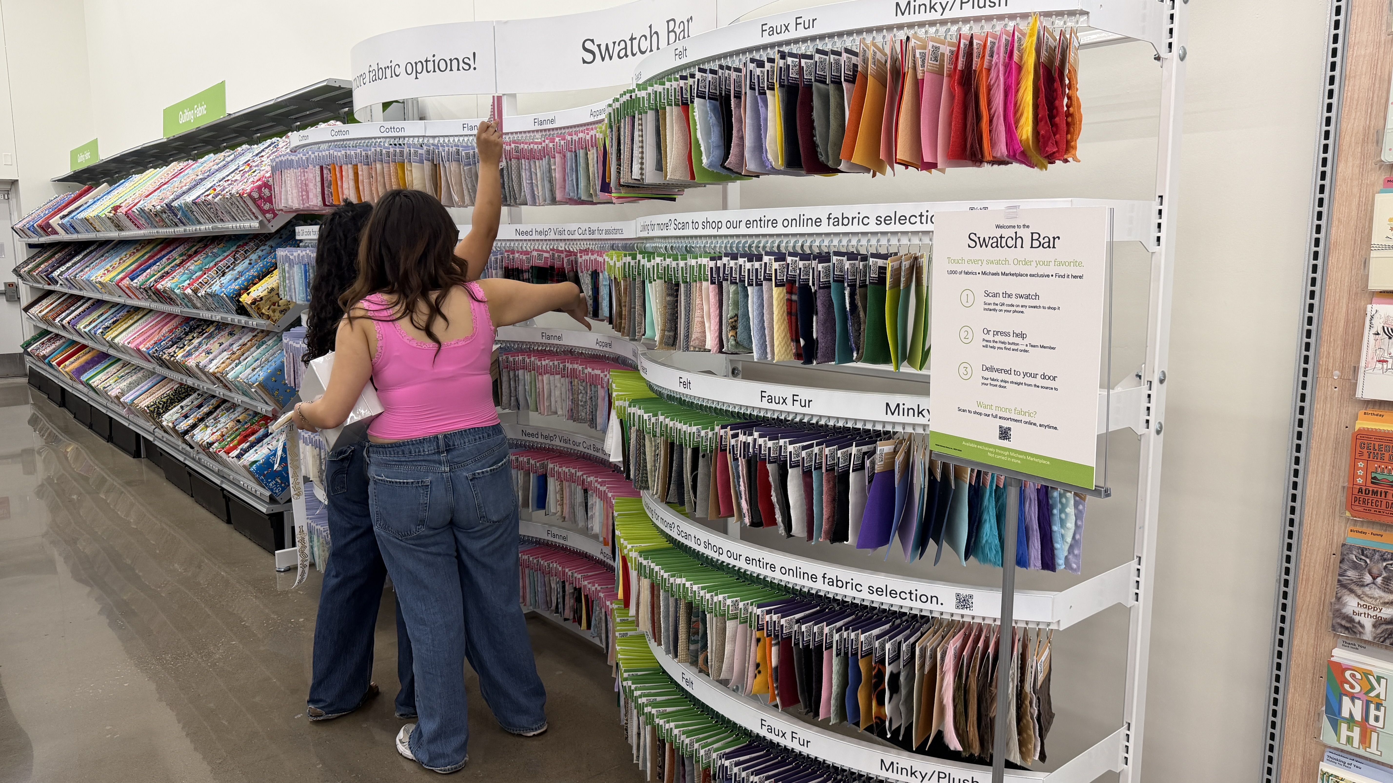 Back view of a shopper in a pink sleeveless top and jeans, reaching for a bright fabric swatch from a curved Swatch Bar display with rows of colorful fabric samples in a store.