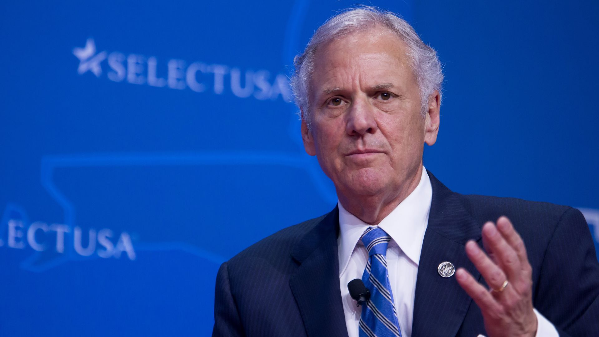 And older adult man gestures while speak on a platform stage, a blue SelectUSA pattern in the background.