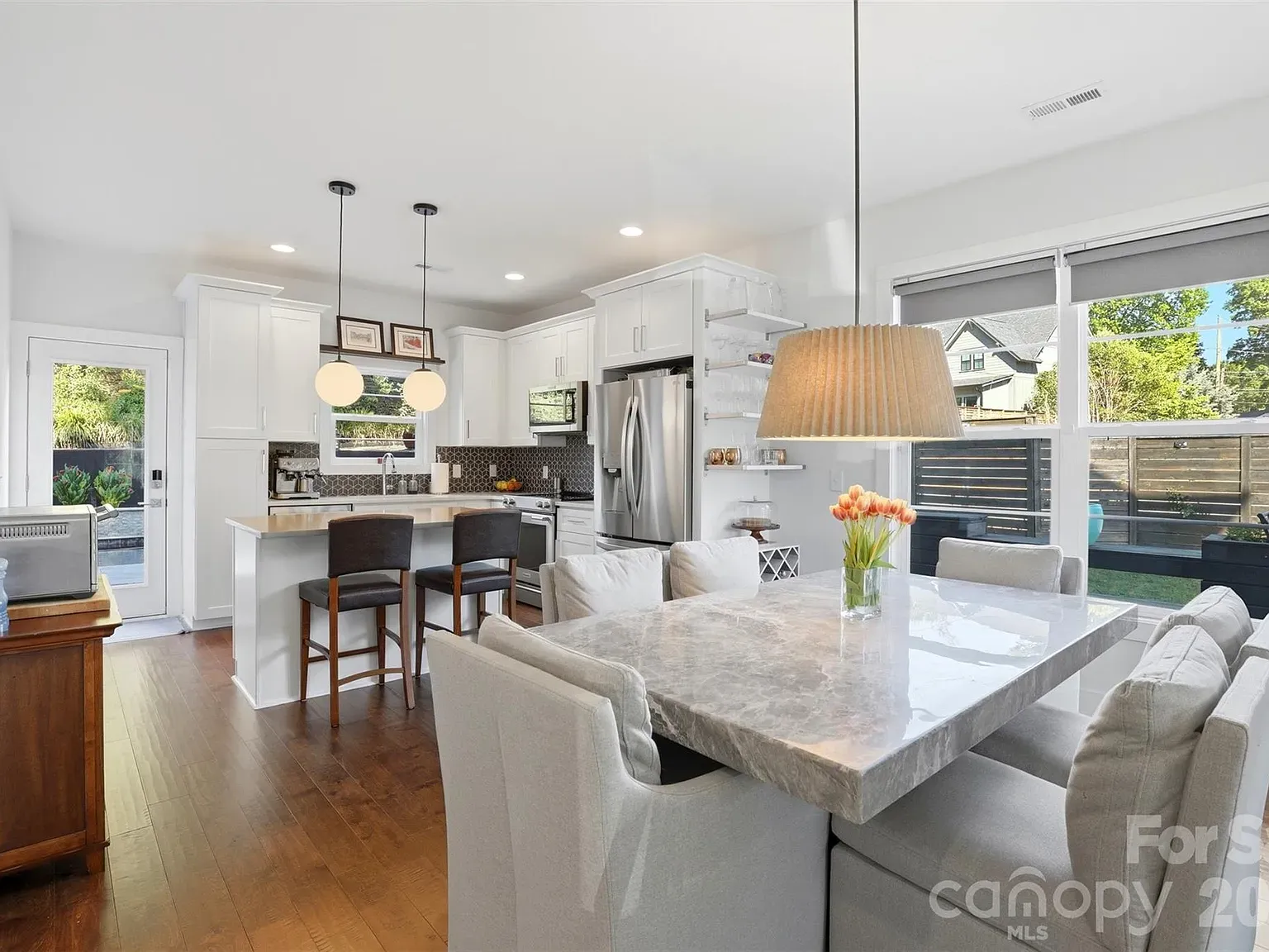 Bright modern kitchen and dining area with white cabinets, stainless steel fridge, marble island with two stools, a marble dining table, large windows, and a vase of orange tulips.