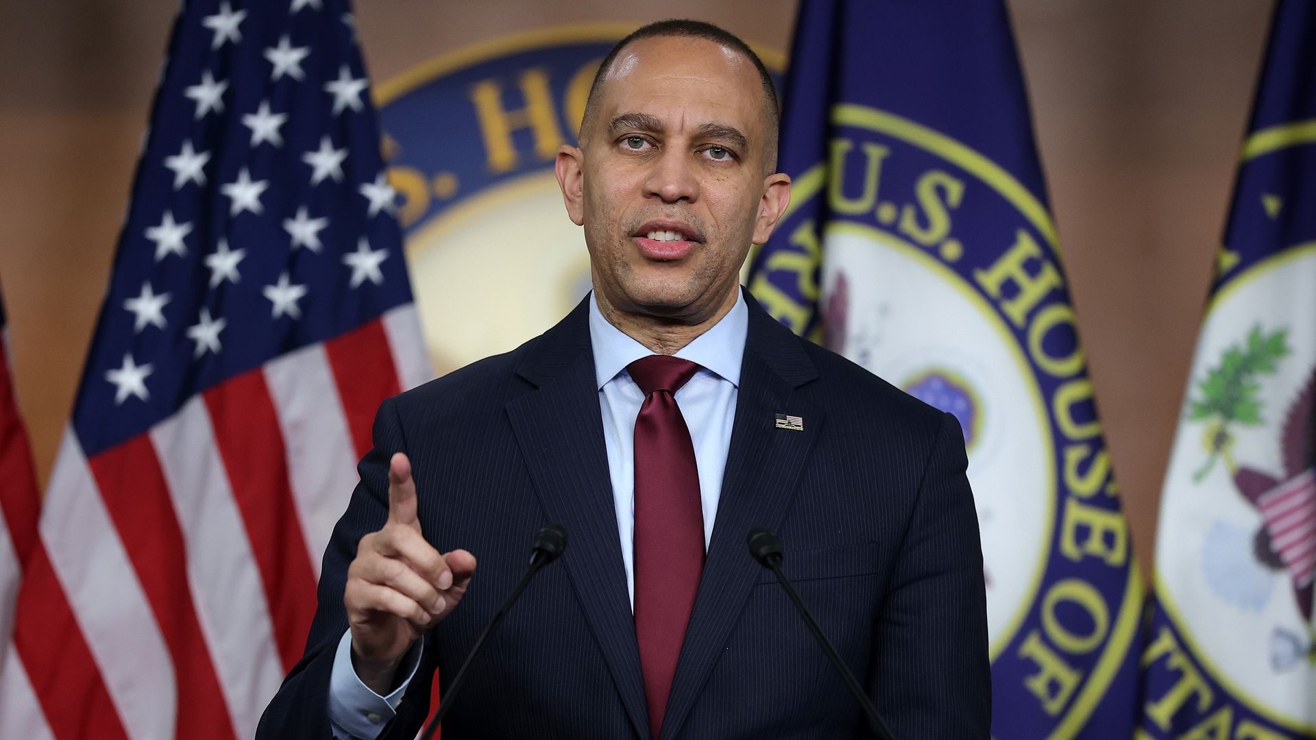 Man in dark suit, white shirt, and red tie speaking at podium with U.S. flag and U.S. House of Representatives flags in the background.