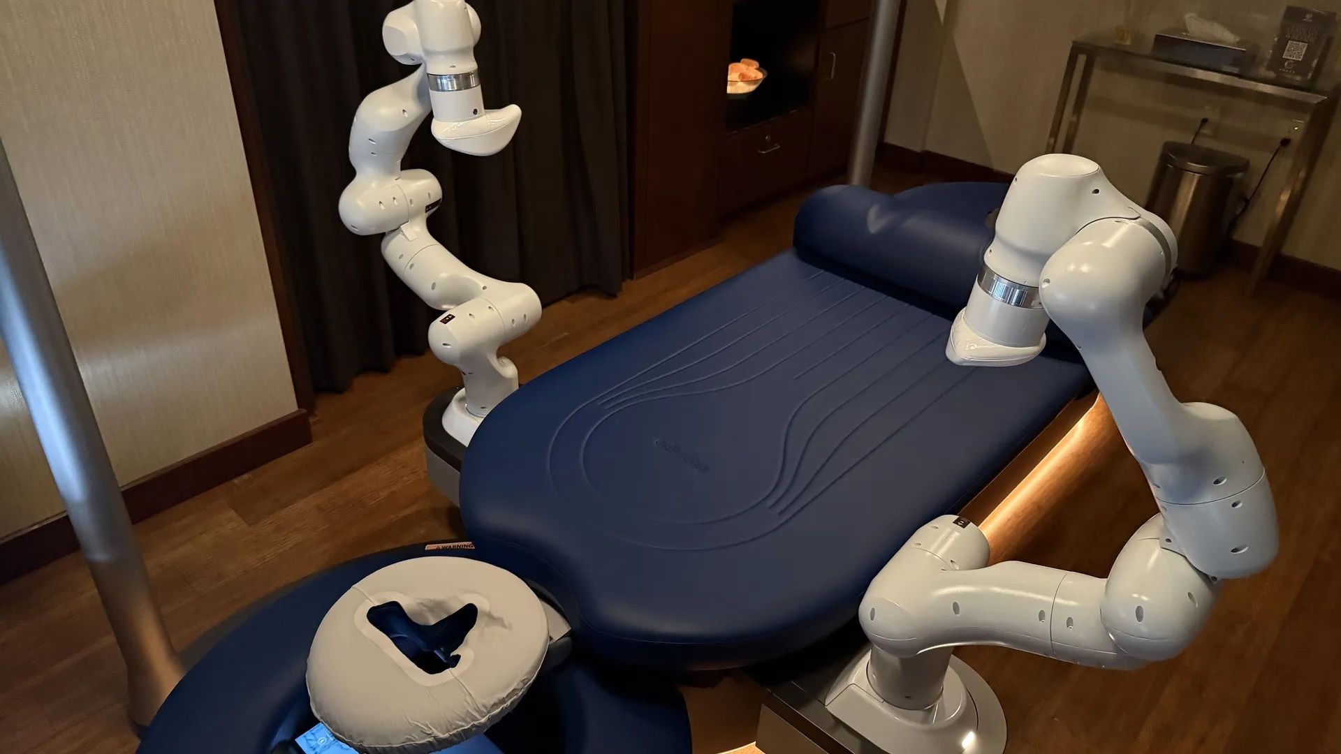 Automated massage bed with white robotic arms and a blue cushion, set in a wooden floor room with dark curtains and a small cabinet in the background.