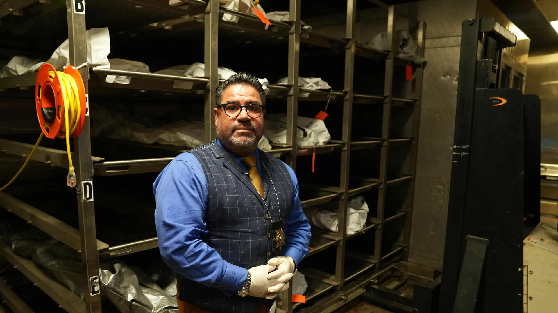 A man wearing white medical gloves stands in front of a row of tall bins with human remains