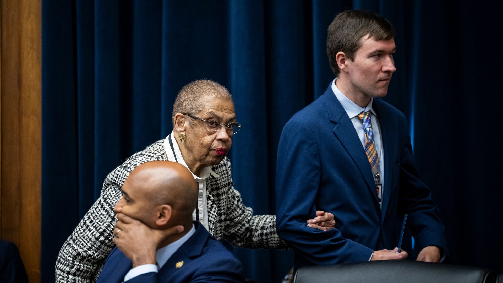 Del. Eleanor Holmes Norton, wearing a black and white pant suit, is escorted by a staffer in a blue suit in front of a blue curtain and behind a wooden dais.