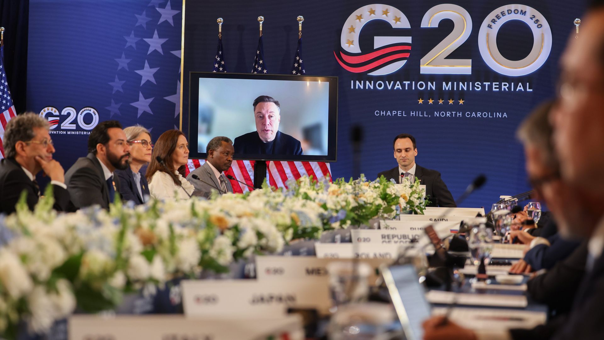 A person on a livestream speaks to a long table in front of a G20 sign