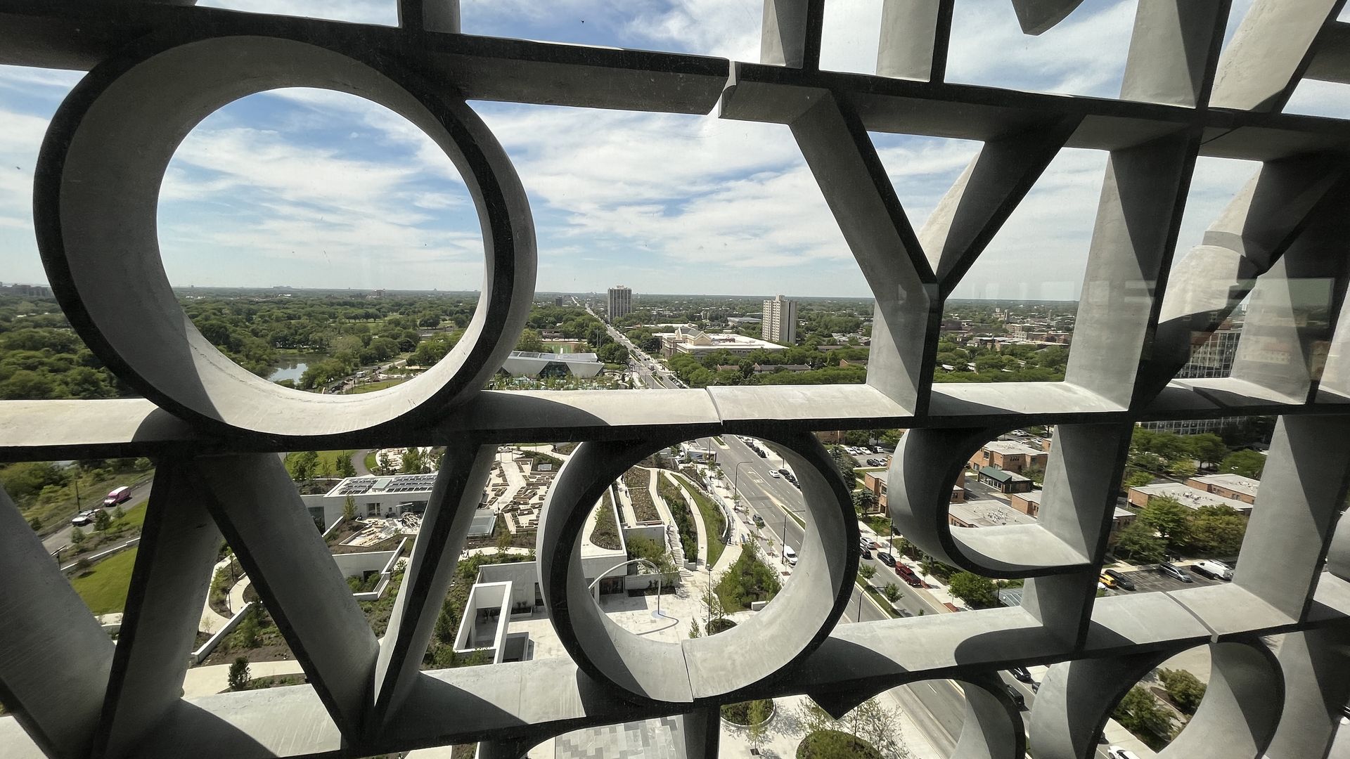 View through a dark geometric metal lattice with circular loops and straight bars, overlooking a city below with roads, buildings, and green trees under a blue sky.
